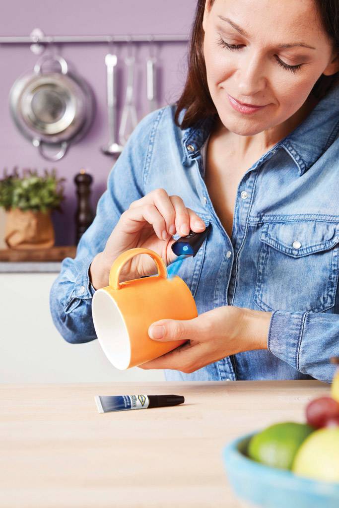 Eine Frau in einer Küche repariert mit Kleber eine orangefarbene Tasse. Im Hintergrund sind Küchenutensilien und eine Schüssel mit Obst.