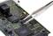 Eine Pinzette hält einen kleinen, schwarzen Chip oberhalb einer grünen Leiterplatte mit zahlreichen elektronischen Bauteilen.