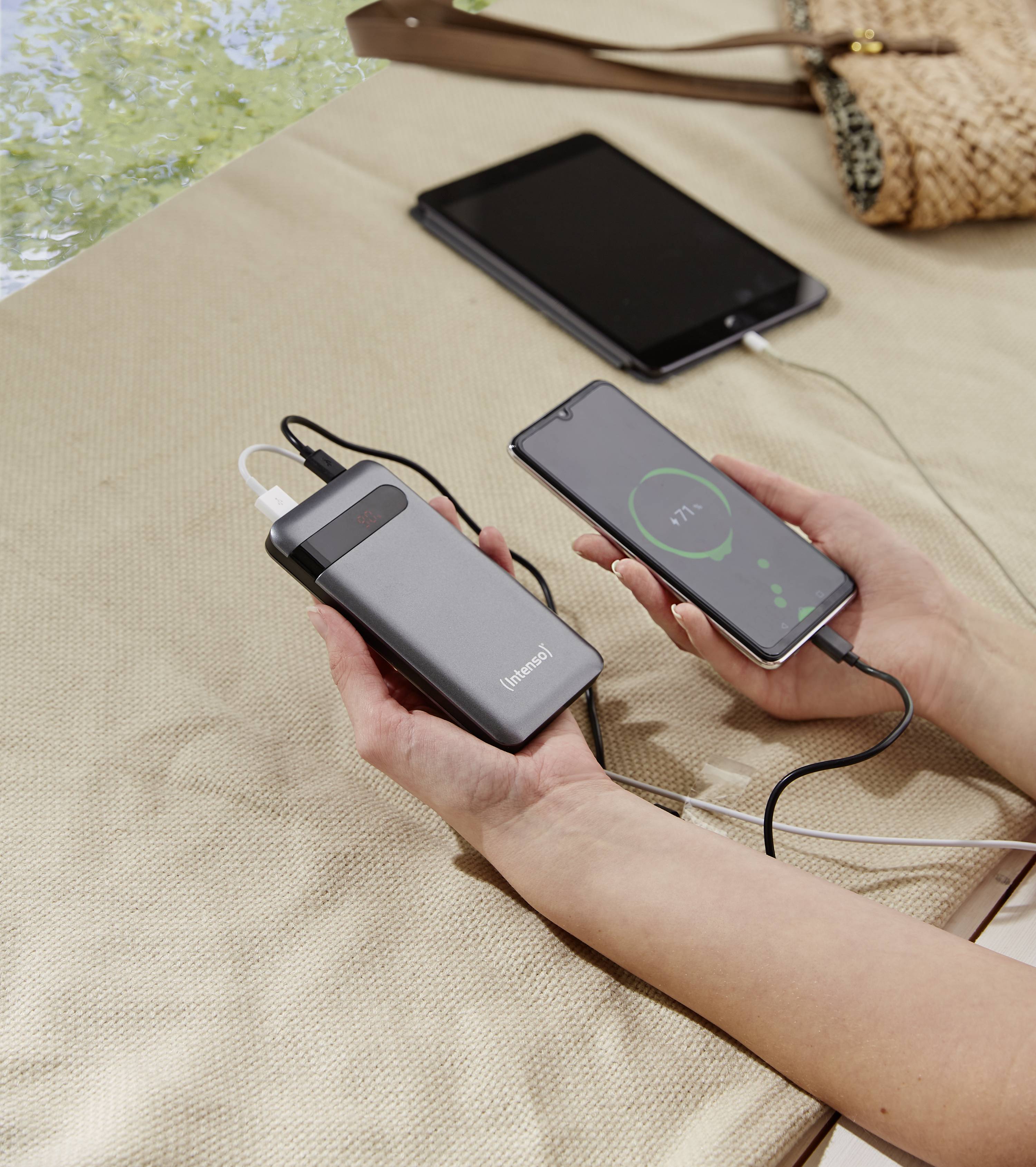 Eine Person hält eine Powerbank, die ein Smartphone auflädt. Das Smartphone zeigt 71% Ladestand an. Im Hintergrund liegt ein Tablet auf dem Tisch.