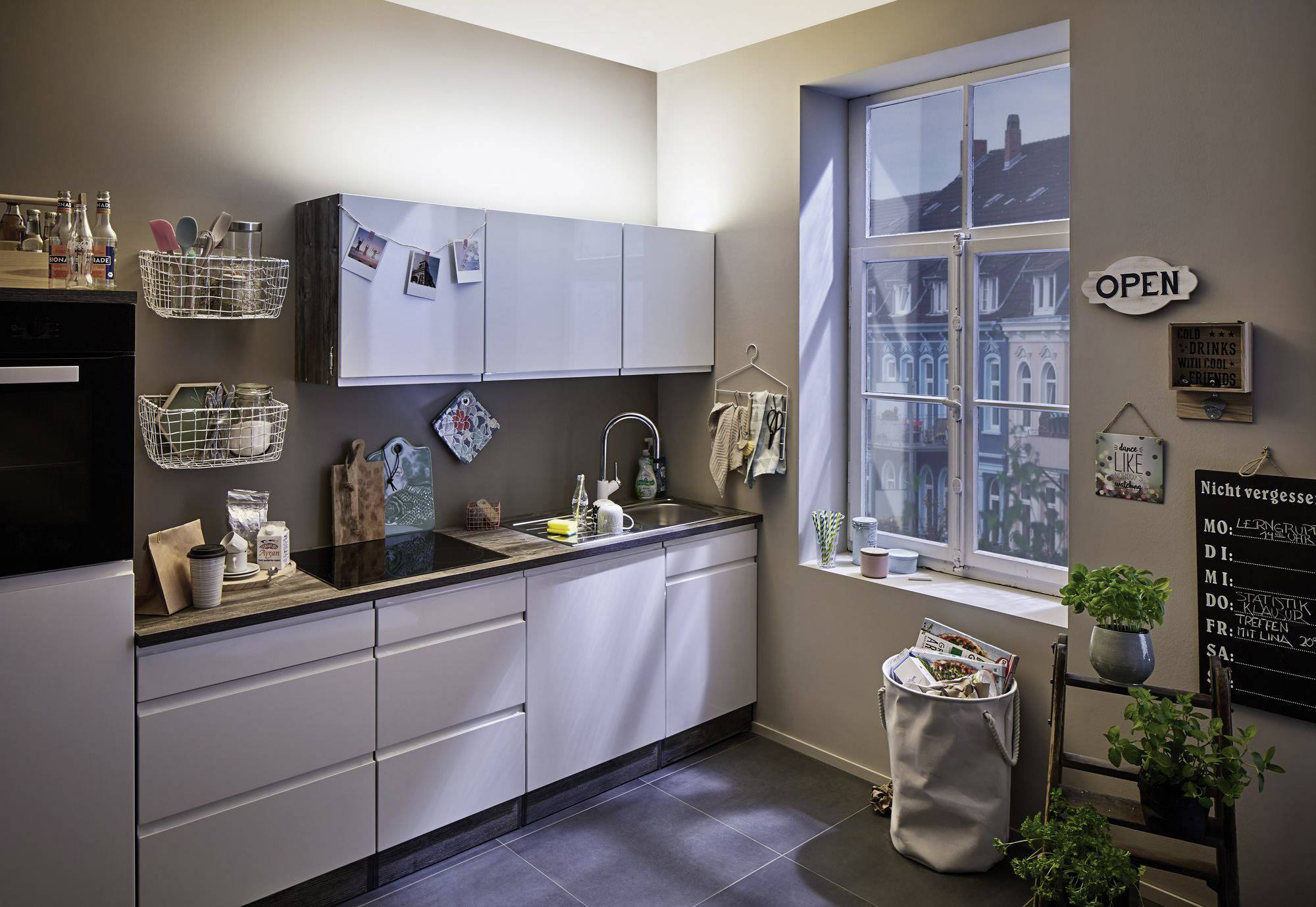 Moderne Küche mit weißen Schränken, einem großen Fenster und dekorativen Elementen wie Pflanzen und Kreidetafel im Hintergrund.