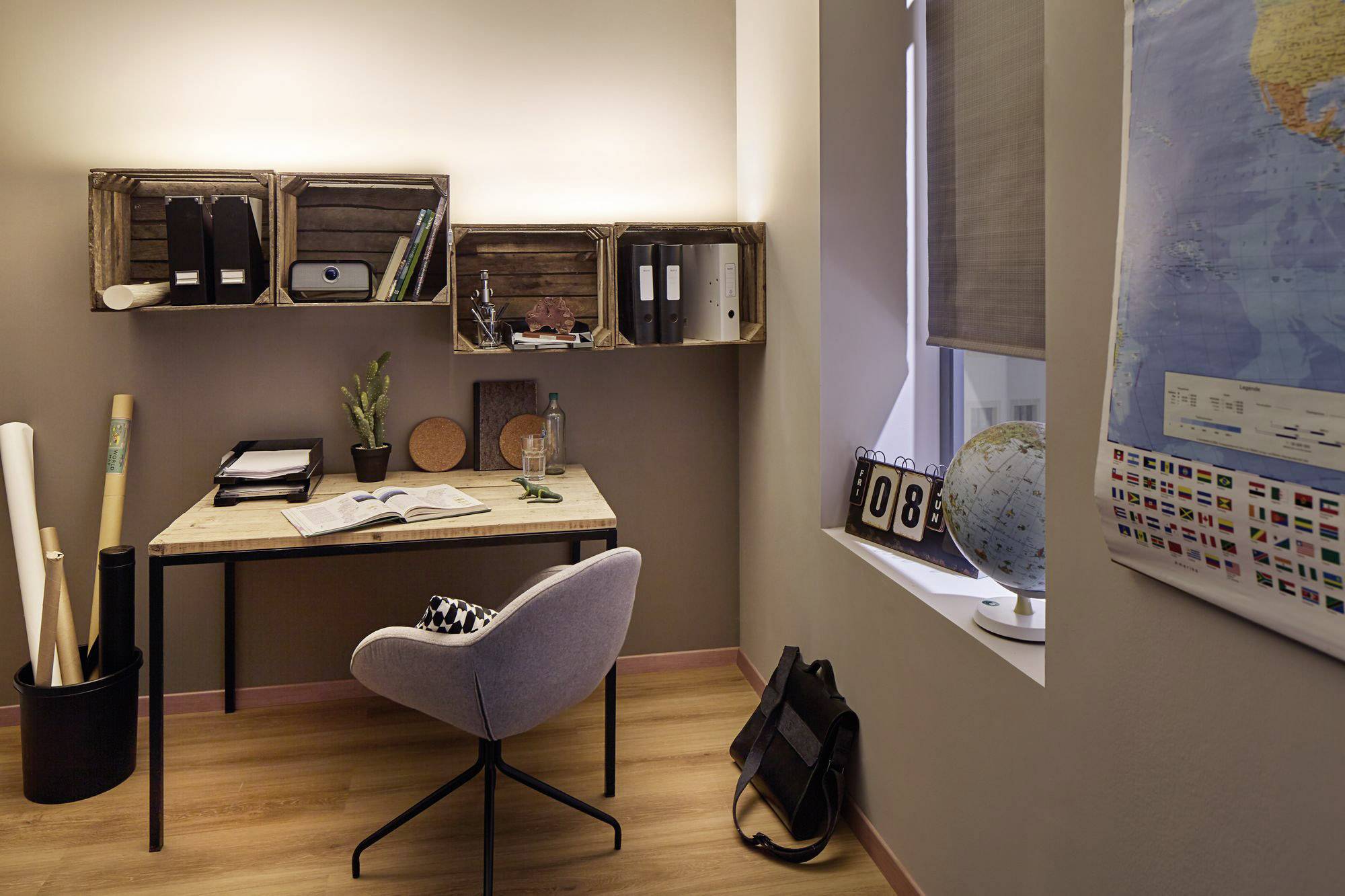 Ein Homeoffice mit einem Schreibtisch, einem Stuhl, Regalboxen an der Wand, Pflanzen und Dekoration. Ein Globus und Kalender auf der Fensterbank.