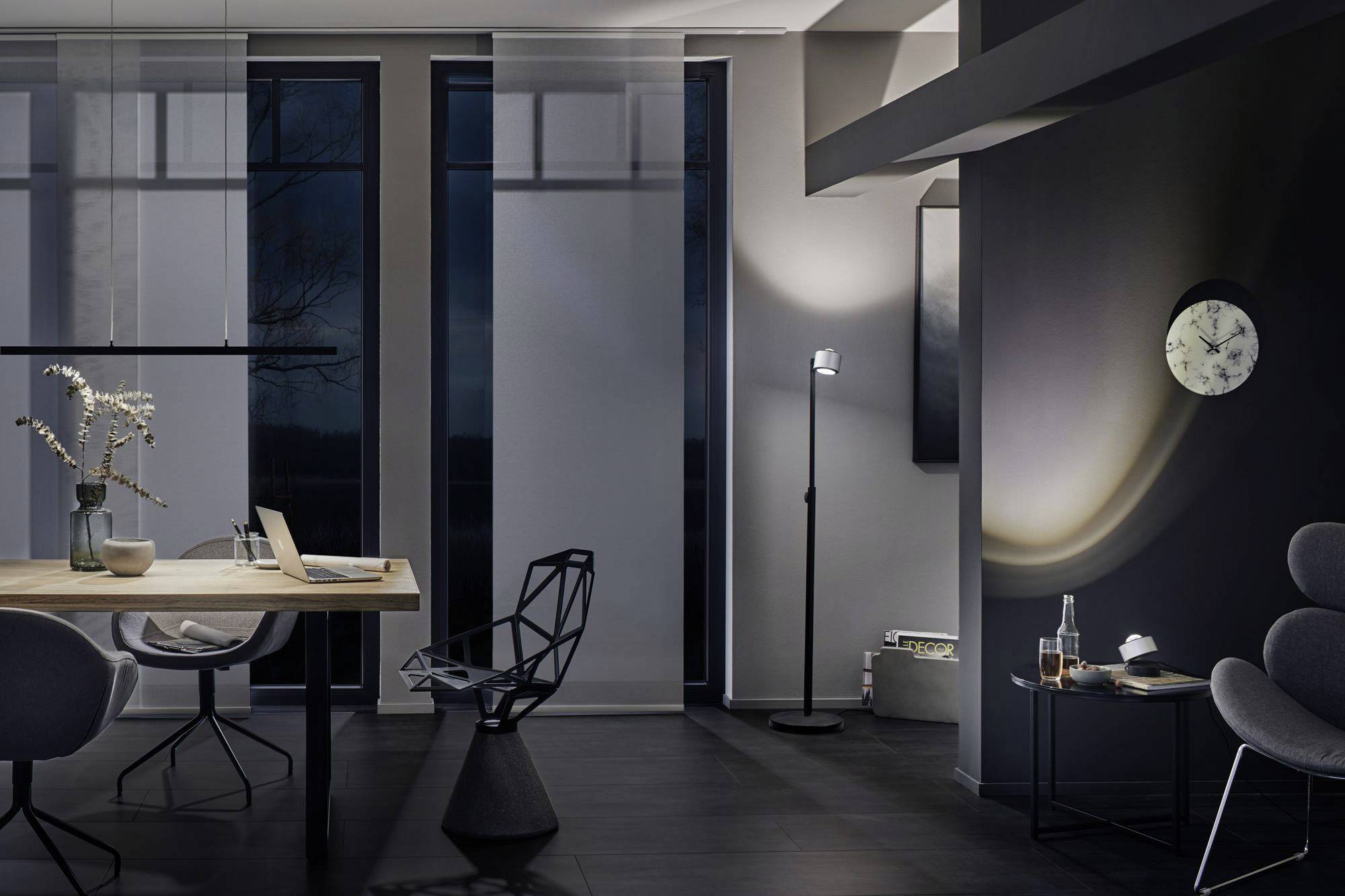 Modern eingerichteter Raum mit großem Tisch, stilvollem Stuhl und Stehlampe. An der Wand hängt eine Uhr. Atmosphäre wirkt beruhigend.