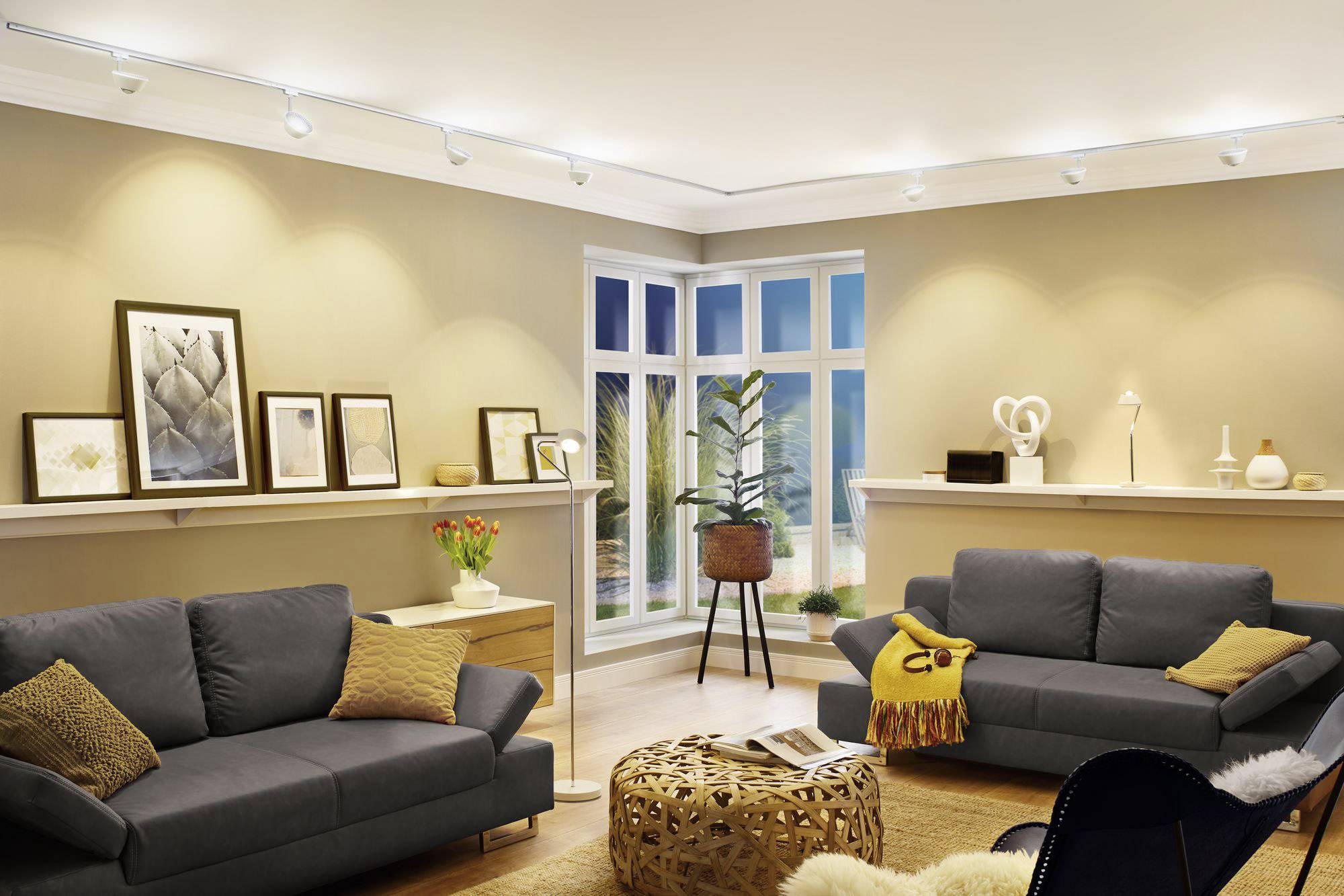 Gemütliches Wohnzimmer mit zwei grauen Sofas, dekorativen Kissen, Kunstwerken an der Wand und Pflanzen vor einem großen Fenster.