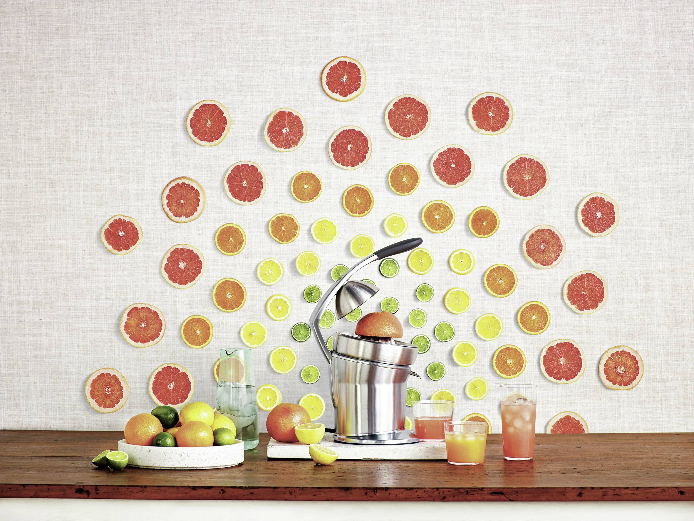 Zitruspresse auf Holztheke mit verschiedenen Zitrusfrüchten, darunter Grapefruits, Zitronen und Limetten; Säfte in Gläsern davor.