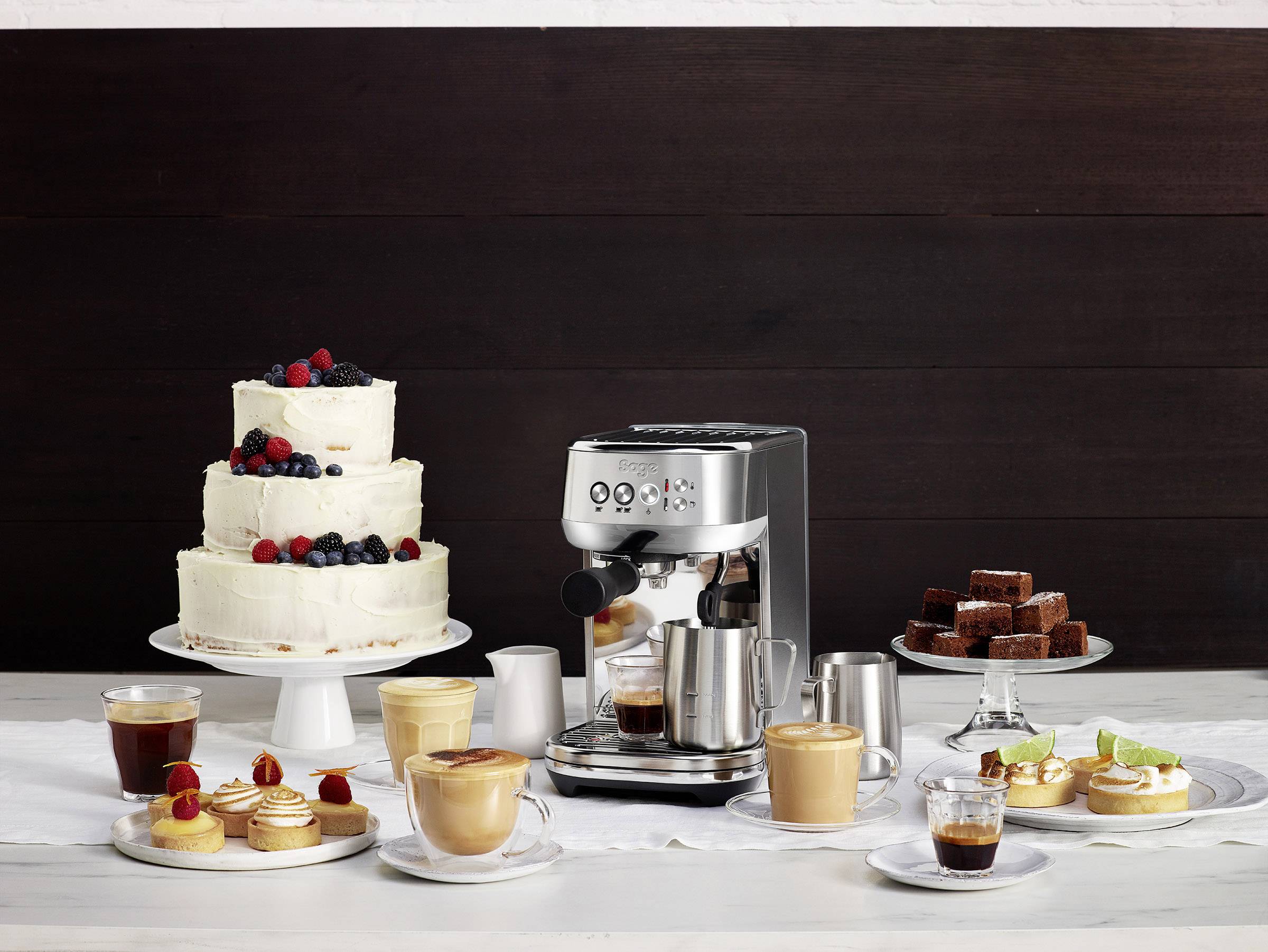Espressomaschine mit Cappuccinos, Kuchen und Gebäck auf einem Tisch. Zwei Torten, kleine Schokoladenstücke und diverse Getränke.