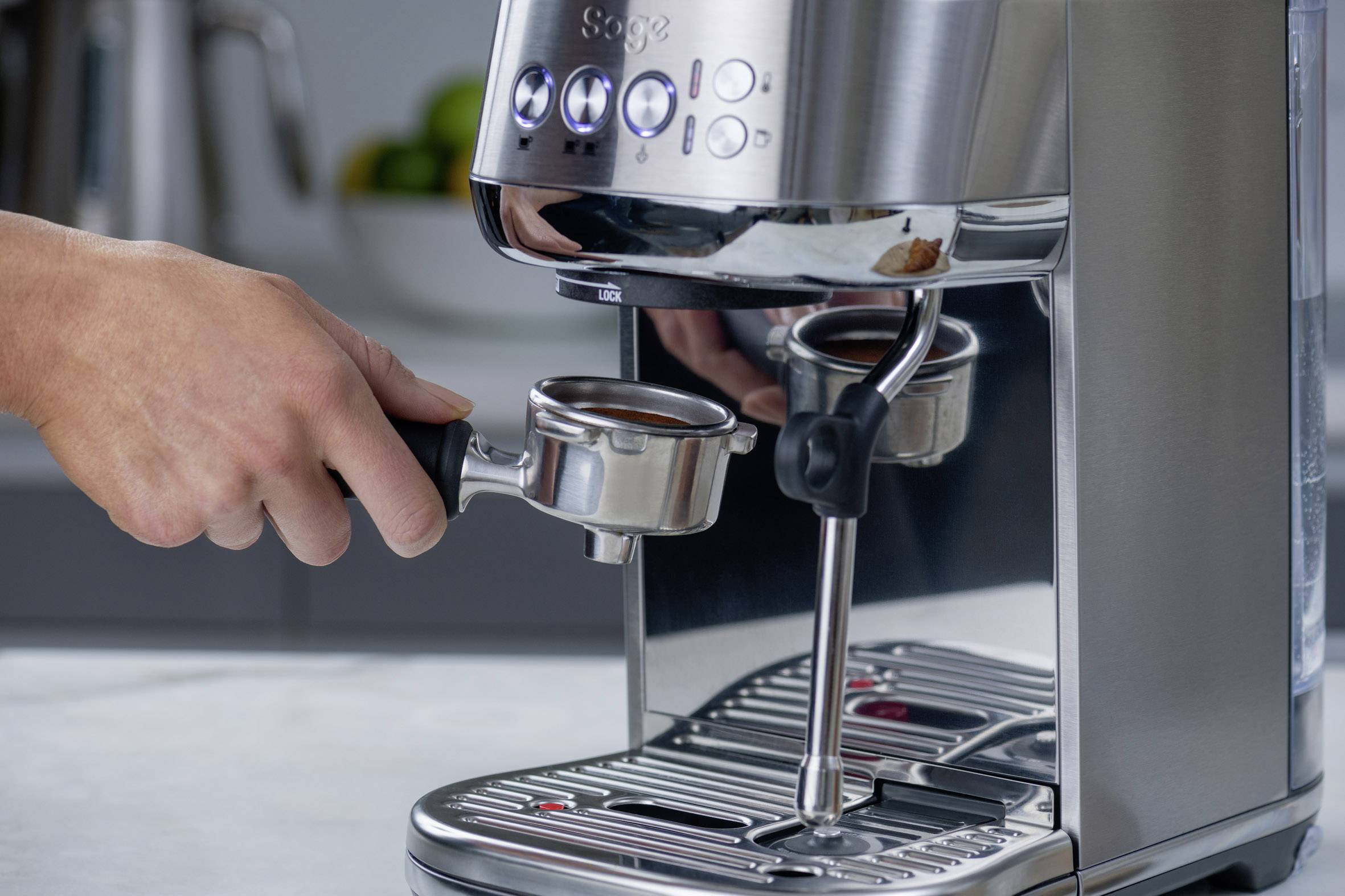 Eine Hand benutzt einen Siebträger an einer Espressomaschine, um frisch gemahlenen Kaffee zuzubereiten. Im Hintergrund sind verschwommene Küchenutensilien erkennbar.