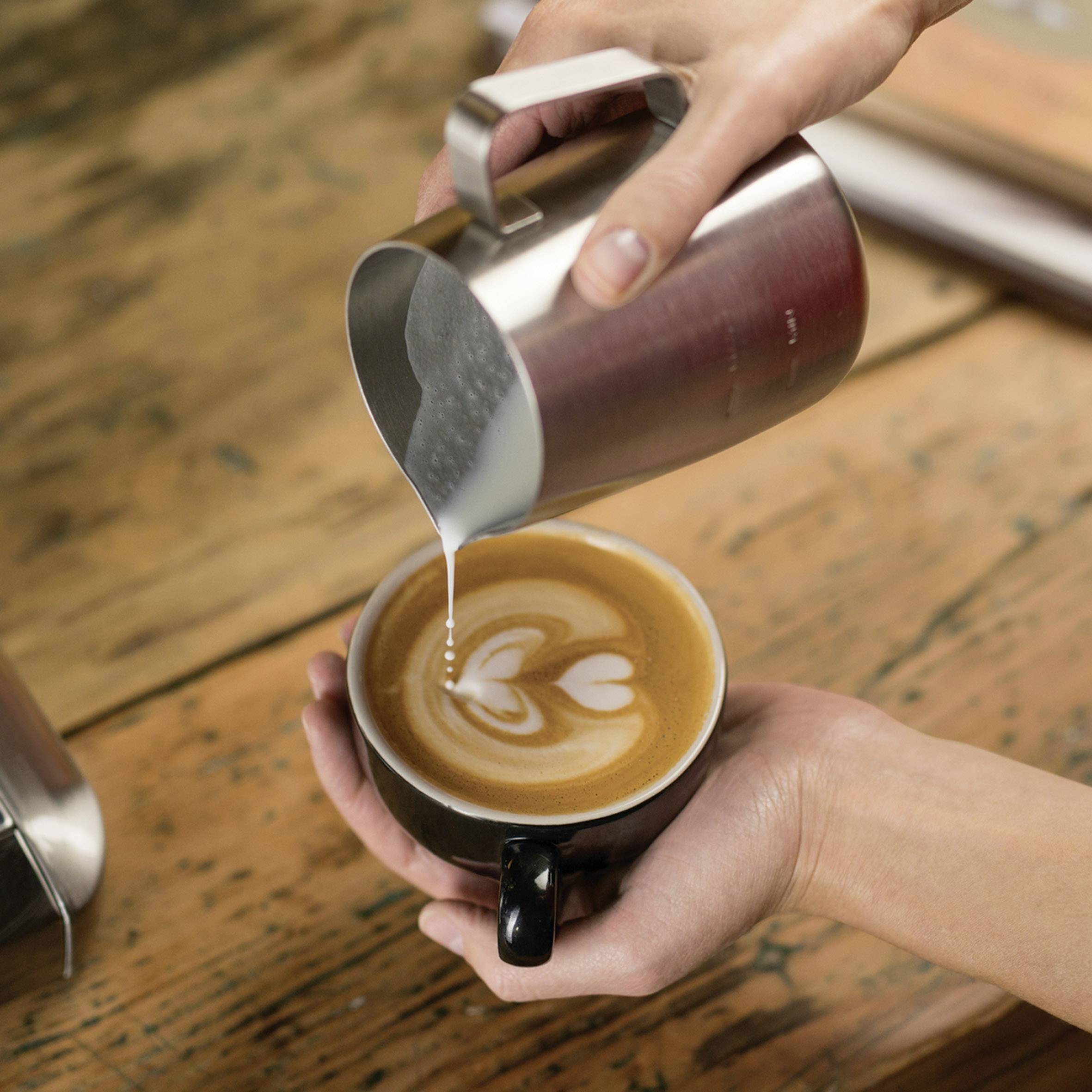 Zwei Hände halten Tassen bei der Latte-Art-Zubereitung, es entsteht ein Herzmotiv aus Milchschaum in einem Cappuccino.