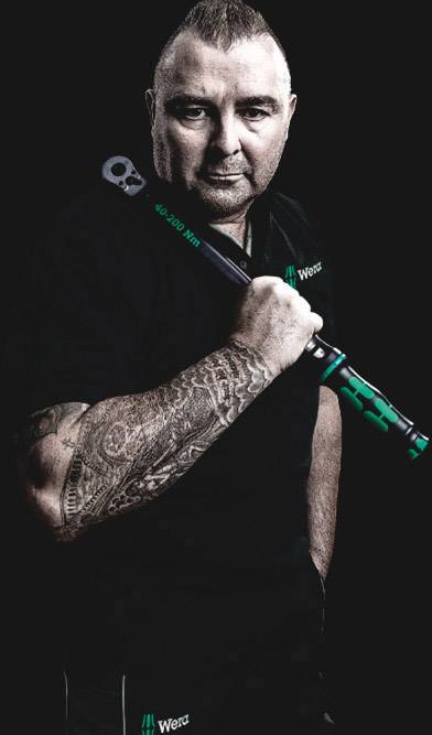 Eine Person mit einem Tätowierten Arm hält einen großen Schraubenschlüssel. Die Person trägt ein schwarzes Hemd mit dem Logo 'Wera'.