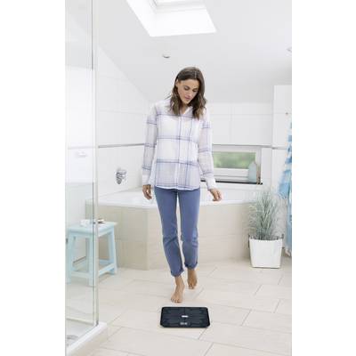 Frau steht barfuß auf einer digitalen Waage in einem modernen Badezimmer. Sie trägt bequeme Kleidung und wirkt konzentriert.