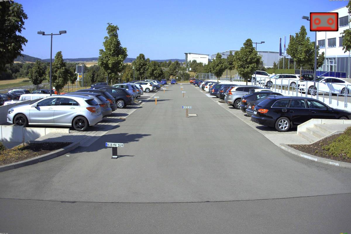 Ein Parkplatz mit mehreren Reihen geparkter Autos. Ein Gebäude im Hintergrund und vereinzelte Bäume an den Seiten.