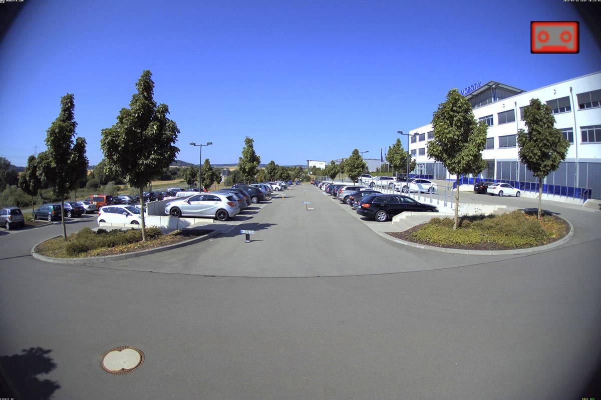 Parkplatz mit Bäumen gesäumt, voll mit geparkten Autos an sonnigem Tag. Bürogebäude rechts im Bild. Klarer blauer Himmel im Hintergrund.