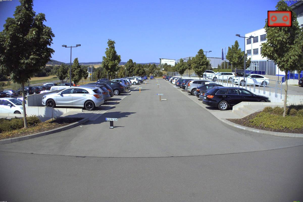 Ein Parkplatz mit Autos auf beiden Seiten, umgeben von Bäumen und Bürogebäuden im Hintergrund. Klare, blaue Himmel und sonniges Wetter.