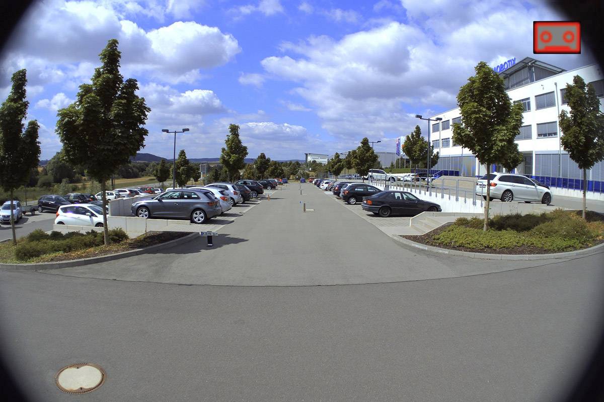 Gebäude mit Parkplatz im Vordergrund, mehrere Autos geparkt. Himmel mit einigen Wolken darüber.