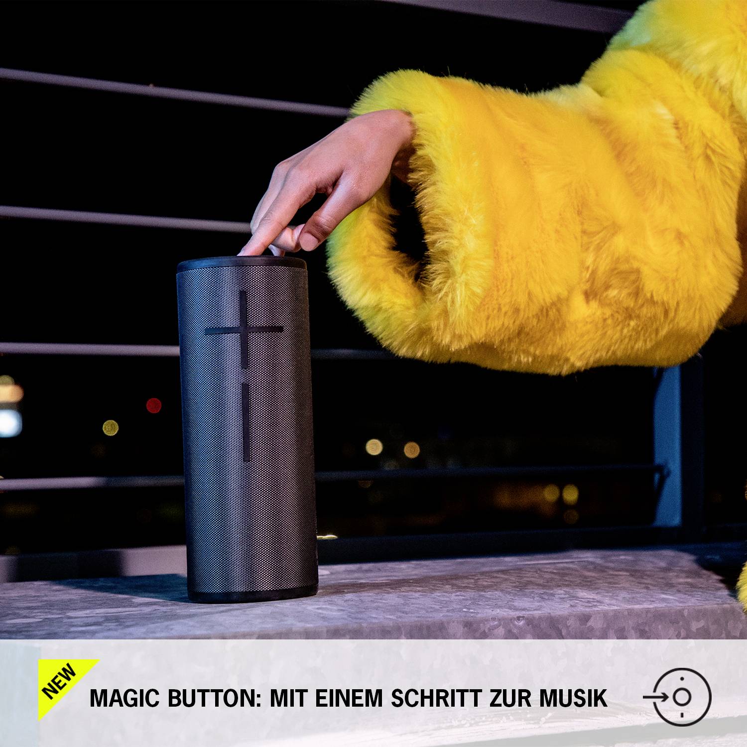 Eine Person mit gelber Jacke drückt einen Knopf auf einem schwarzen, zylinderförmigen Lautsprecher. Text: 'Magic Button: Mit einem Schritt zur Musik'.