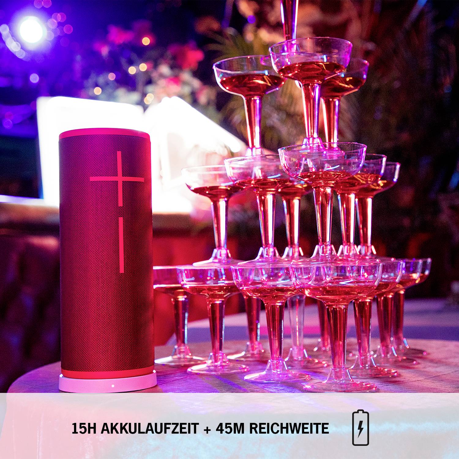 Ein roter Lautsprecher steht neben einem Turm aus gefüllten Champagnergläsern. Text im Bild: '15H Akkulaufzeit + 45M Reichweite'.