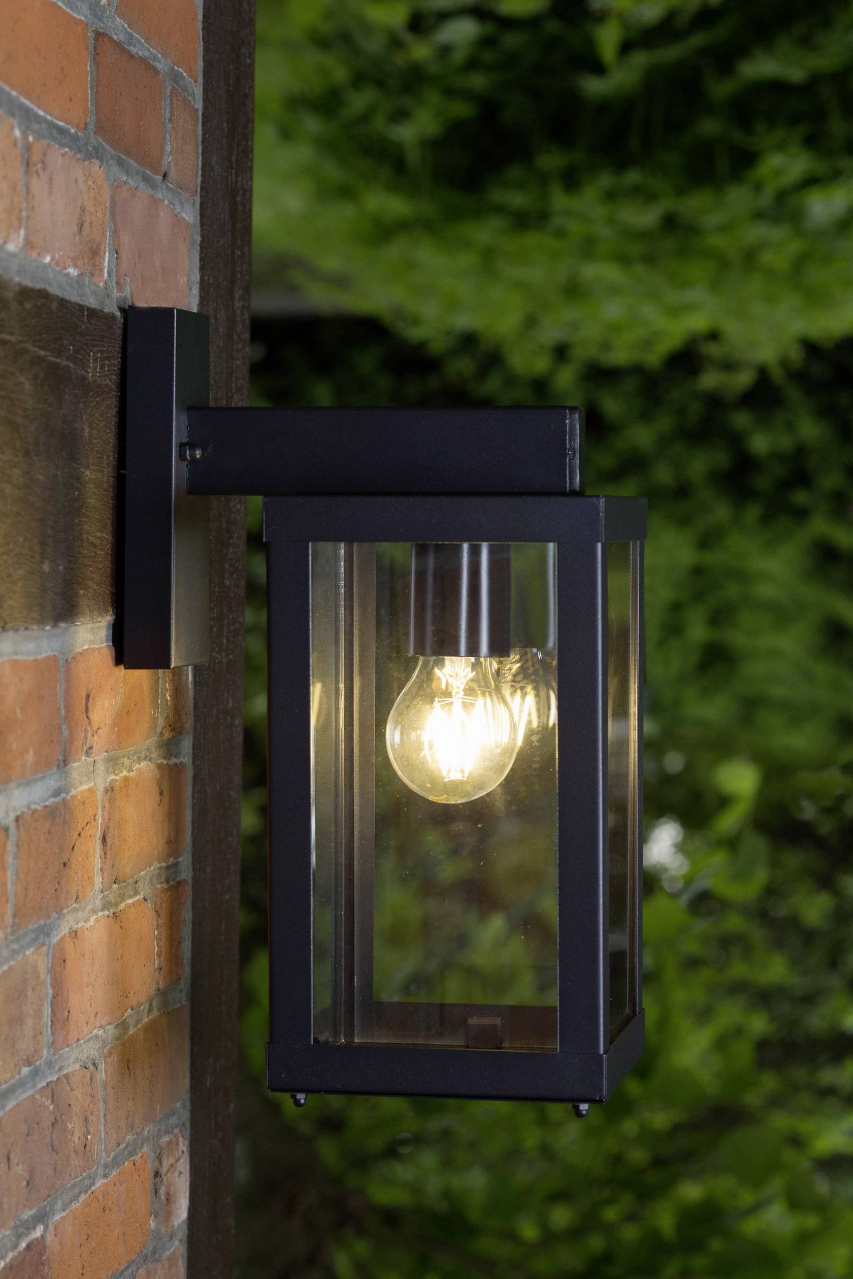 Schwarze Außenlaterne mit sichtbarer Glühbirne an einer roten Backsteinwand befestigt, leuchtet in einem grünen, bewachsenen Gartenbereich.