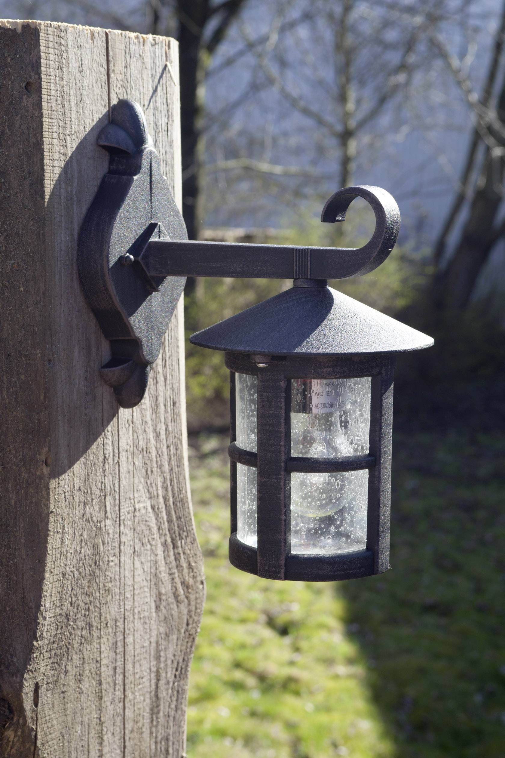 Eine antike Laterne aus schwarzem Metall hängt an einem Holzpfosten im Freien. Im Hintergrund sind Bäume und Gras zu sehen.