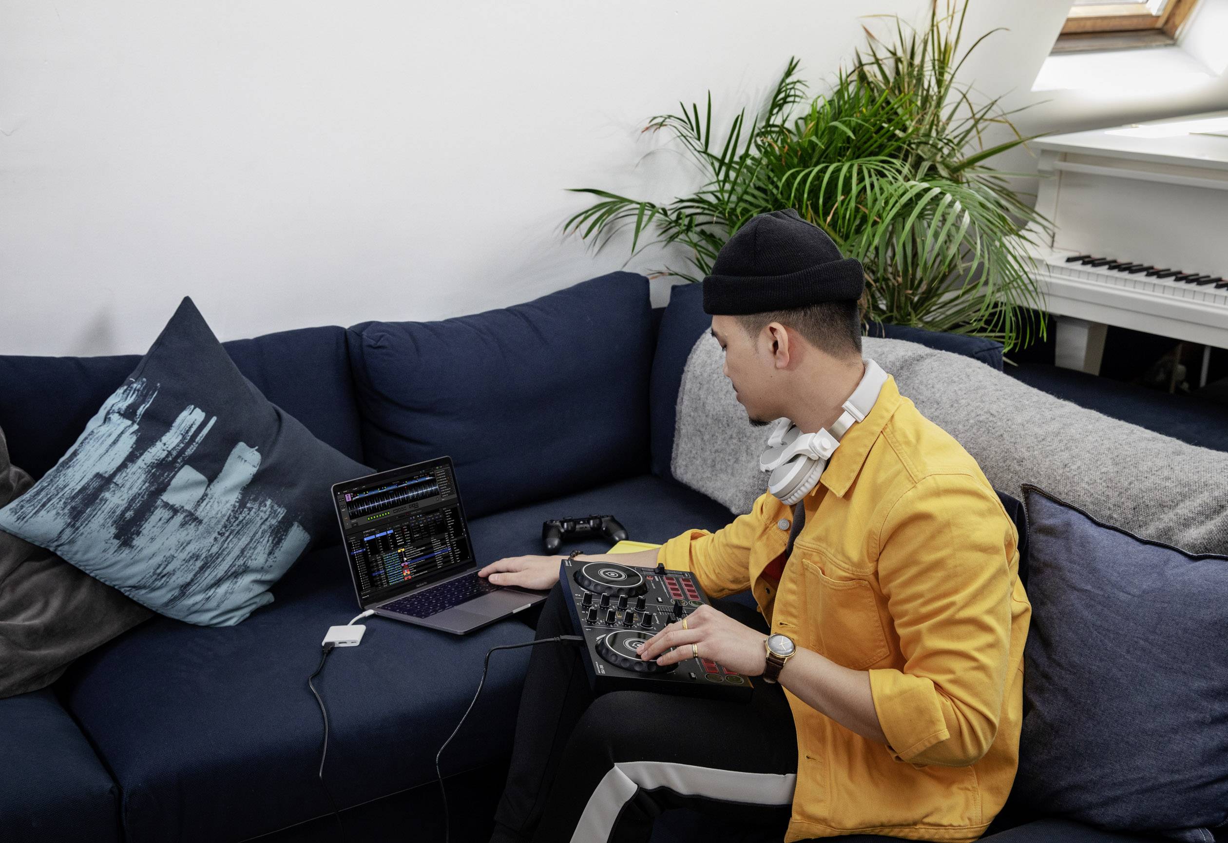 Ein Mann in gelber Jacke mit Kopfhörern mixt Musik auf einem DJ-Controller, der mit einem Laptop auf einem blauen Sofa verbunden ist.