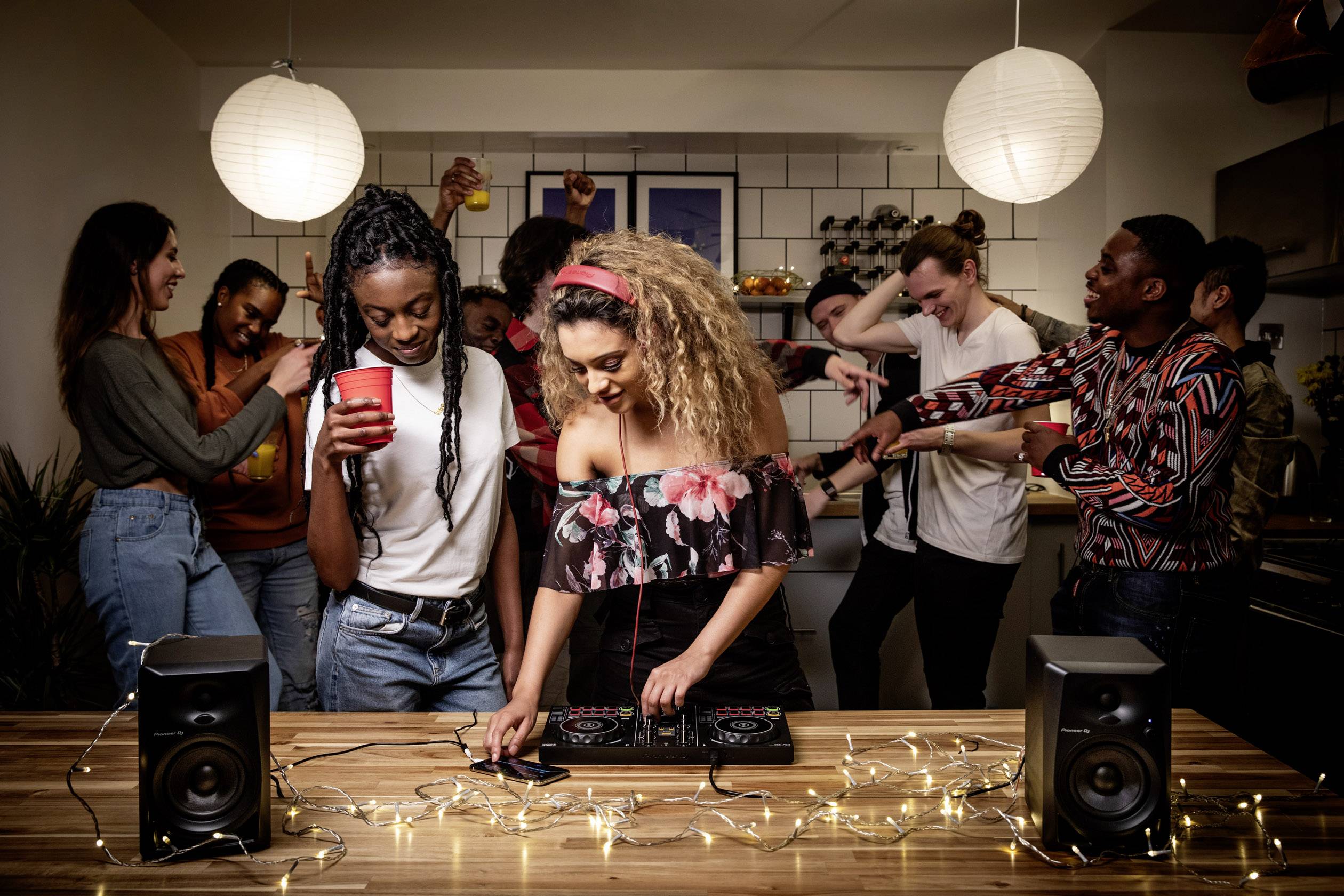 Eine Gruppe junger Erwachsener auf einer Party in einer Küche, mit einer Frau, die Musik mixt, während andere tanzen und plaudern.