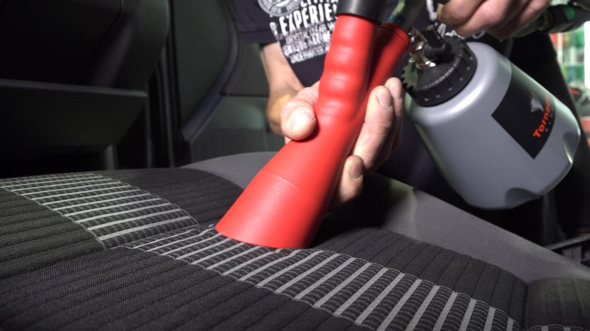 Eine Person reinigt einen Autositz mit einem speziellen Reinigungsgerät. Das Gerät hat eine rote Düse und wird auf den Stoff gehalten.