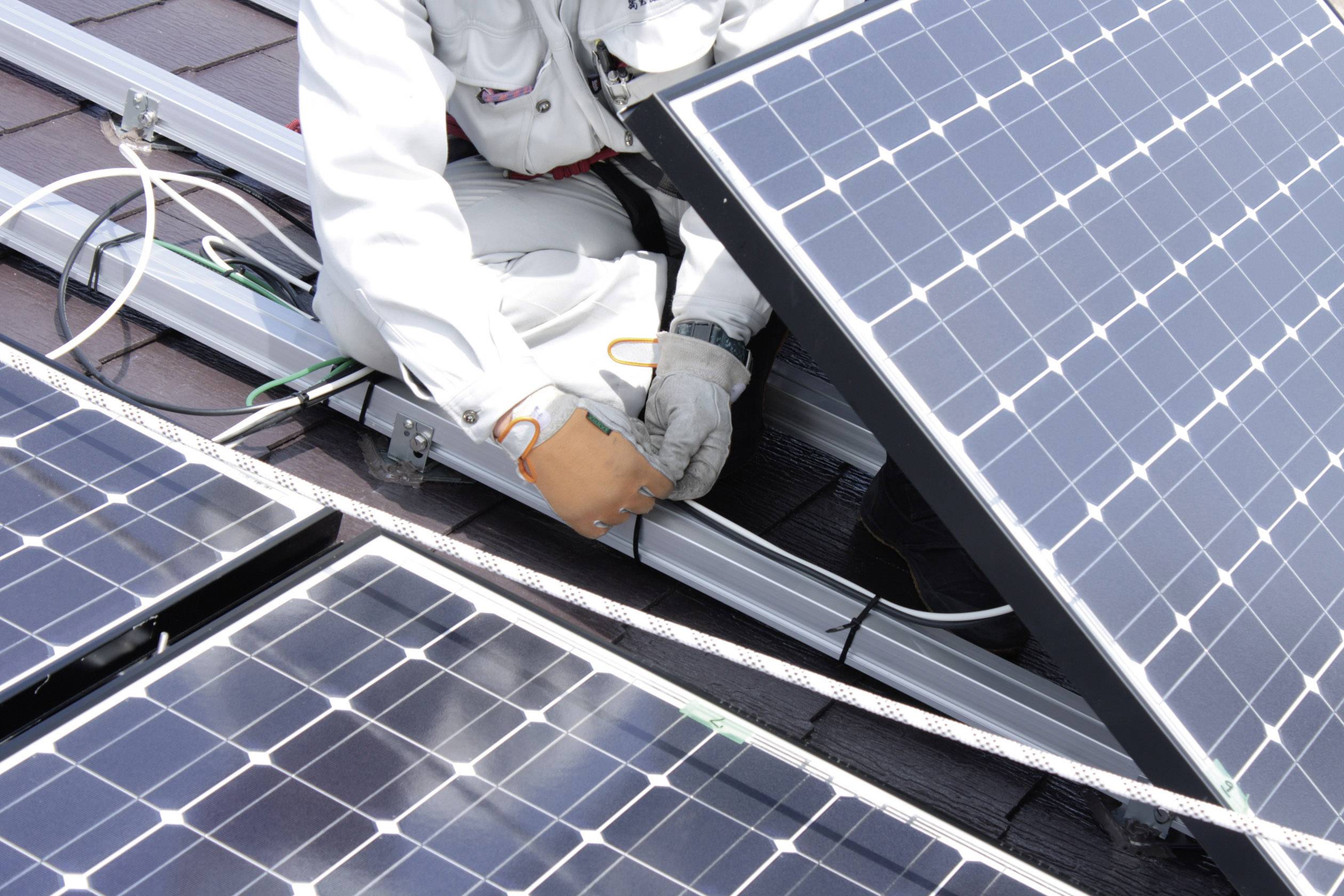 Eine Person installiert Solarmodule auf einem Dach. Sie trägt Schutzkleidung und arbeitet sorgfältig an den elektrischen Verbindungen der Module.