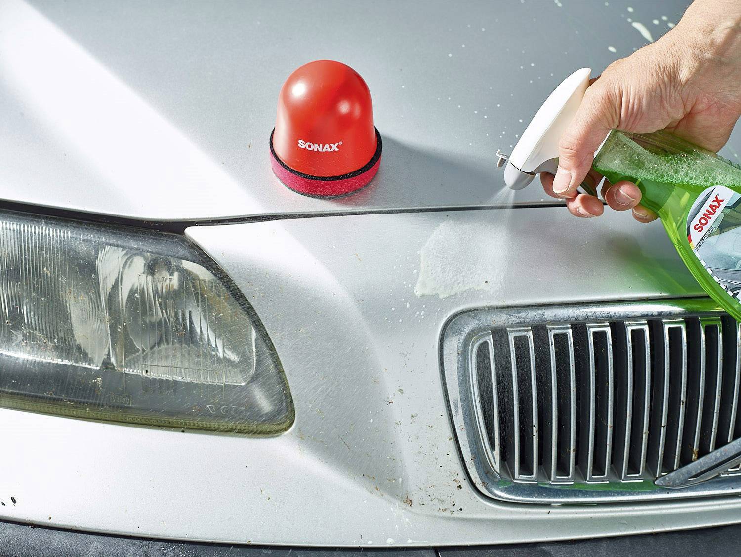 Eine Person reinigt die Motorhaube eines Autos mit einem grünen Reinigungsmittel. Auf der Haube steht ein rotes SONAX-Produkt.