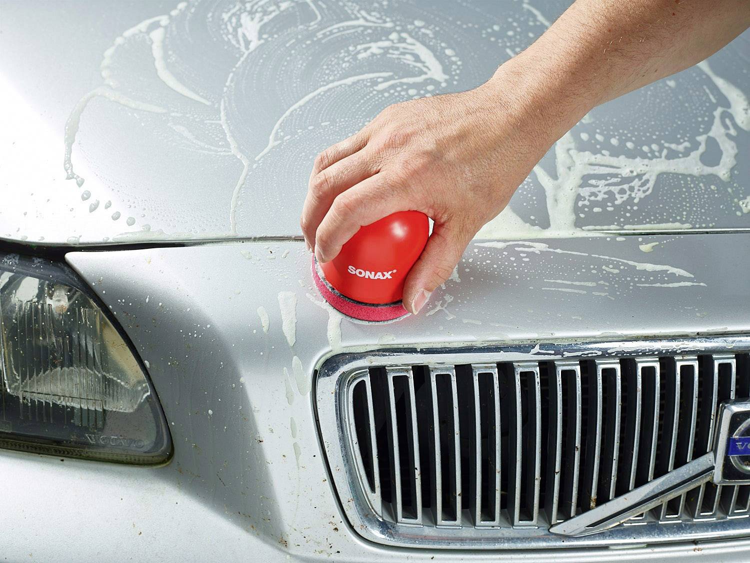 Eine Hand poliert die Motorhaube eines Autos mit einer roten Polierpaste von Sonax.