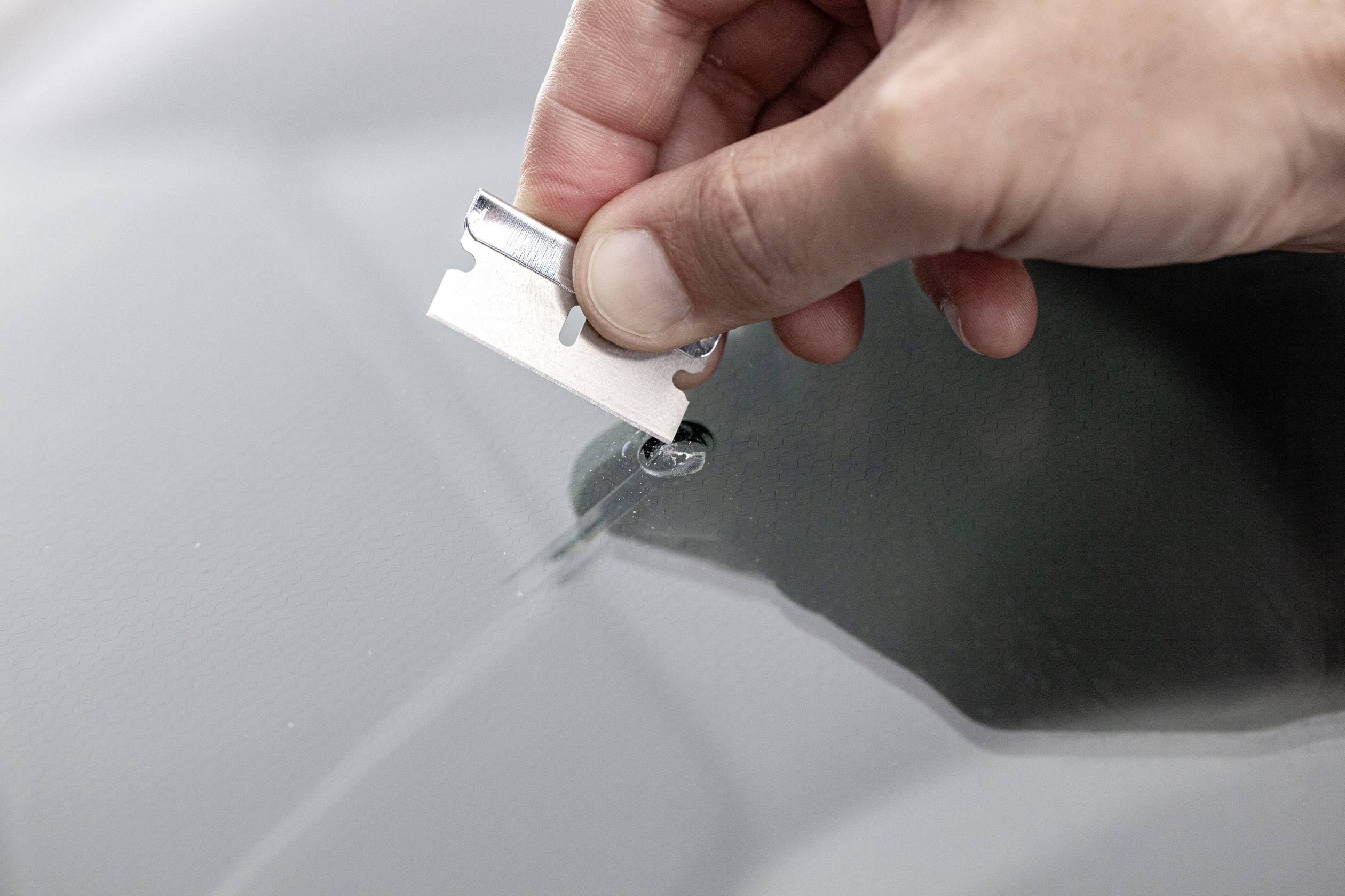 Eine Hand hält eine Rasierklinge über einen Glassplitter auf einer Autowindschutzscheibe und bereitet sich darauf vor, den Splitter zu entfernen.