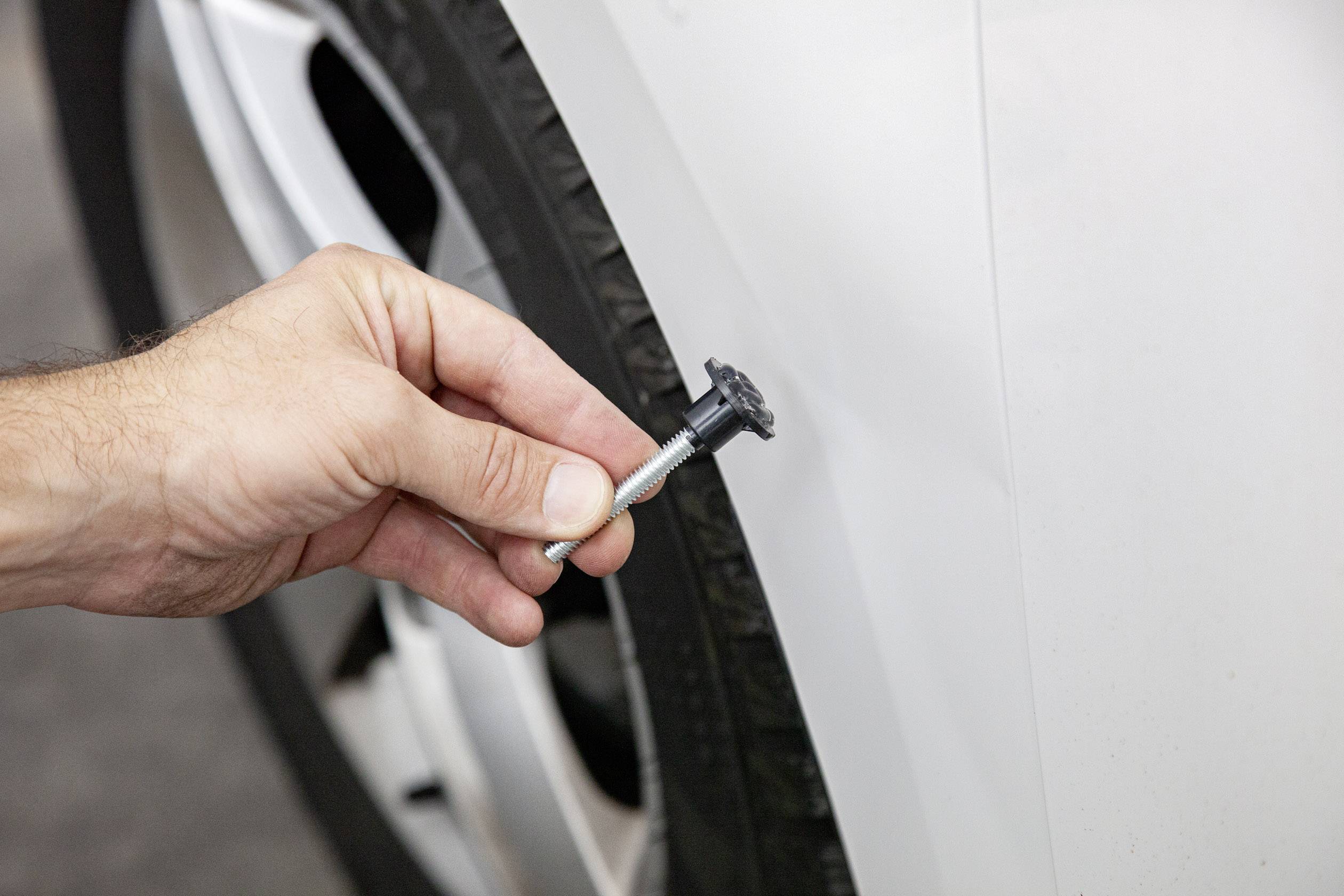 Eine Hand hält einen Schraubenanker nahe einem Autoreifen, während ein Kratzer auf der Karosserie sichtbar ist.