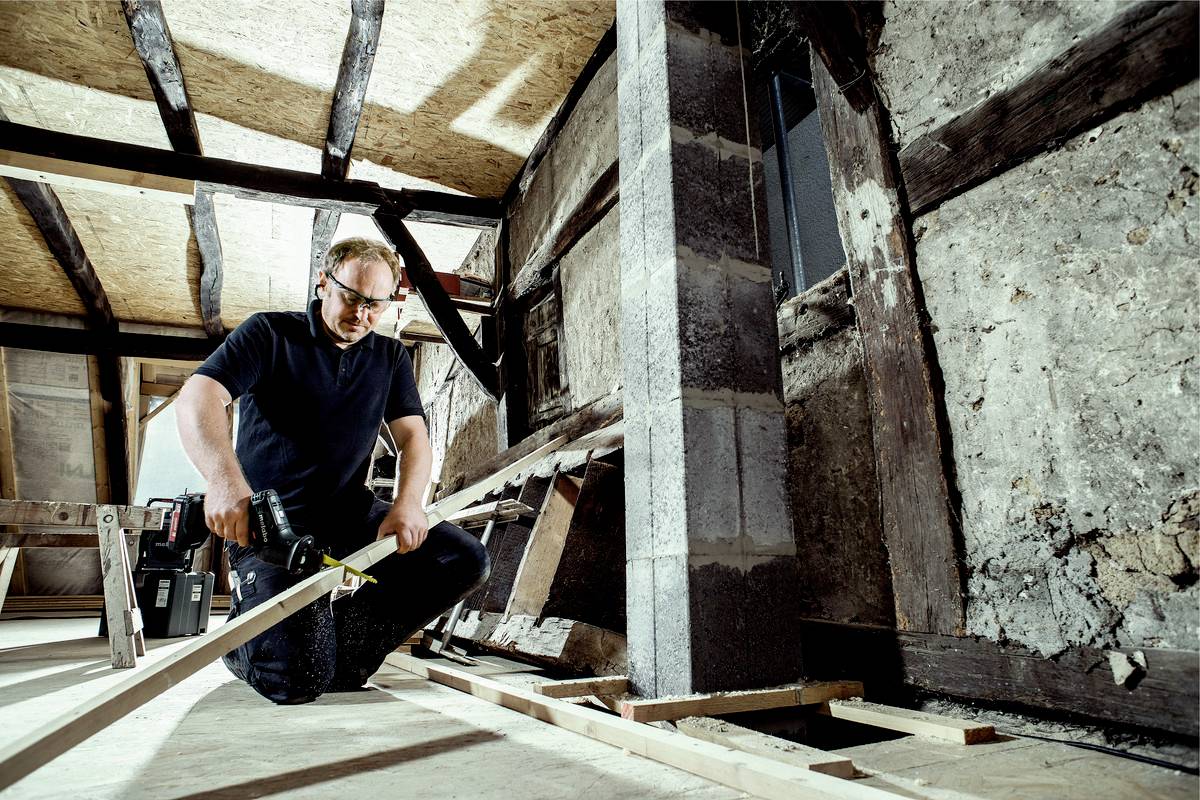Ein Mann arbeitet mit einer Bohrmaschine in einem renovierungsbedürftigen Raum. Er trägt Schutzbrille und umgebende Strukturen sind alt.