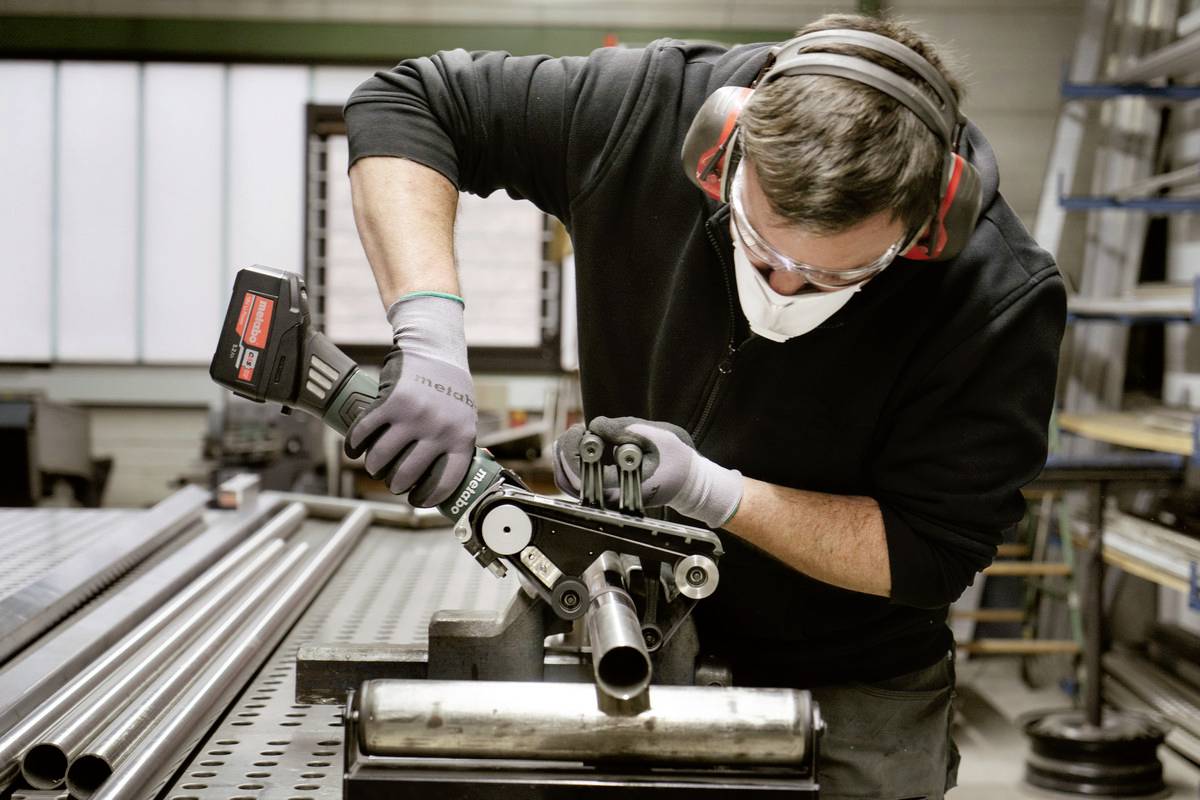 Ein Mann in Schutzkleidung arbeitet an einem Metallrohr mit einem Akkuwerkzeug in einer Werkstatt.