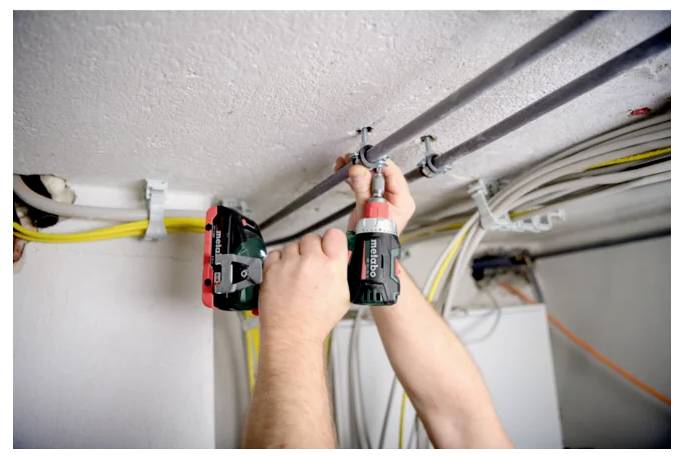 Elektriker, der einen Bohrer verwendet, um eine Leitung an der Decke zu befestigen, mit verlegten Kabeln in einem unfertigen Raum.