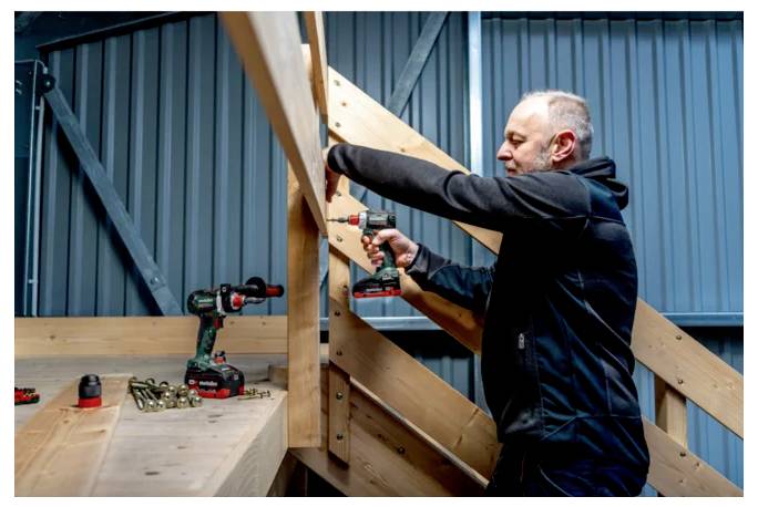 Ein Mann benutzt einen kabellosen Bohrer, um Holzbalken an einem Treppenbauprojekt im Innenbereich zu befestigen.