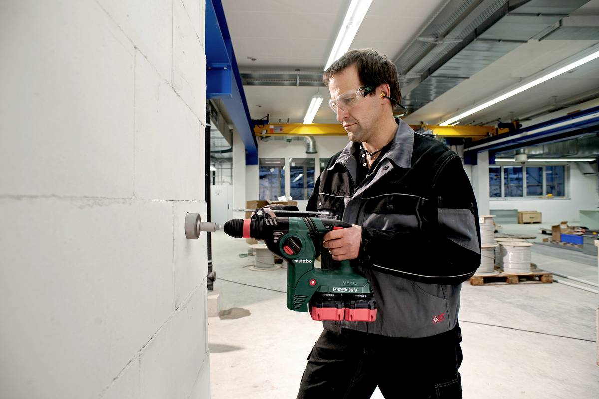 Ein Mann in Schutzbrille und Arbeitskleidung bohrt mit einer grünen Bohrmaschine in eine weiße Wand in einer industriellen Werkstatt.