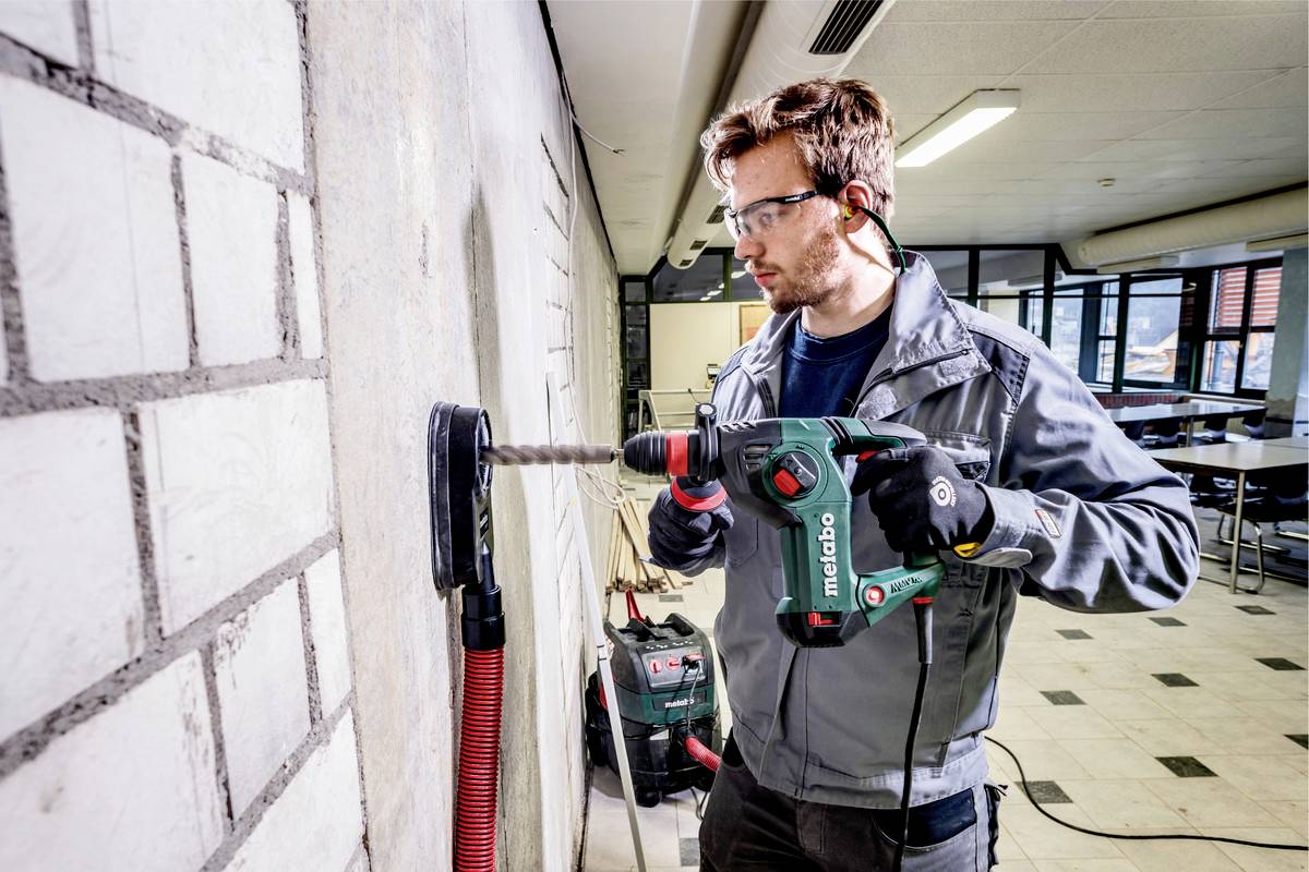 Ein Mann bohrt mit einer grünen Bohrmaschine in eine Wand. Er trägt Schutzbrille und Ohrstöpsel. Im Hintergrund sind Möbel und Fenster.
