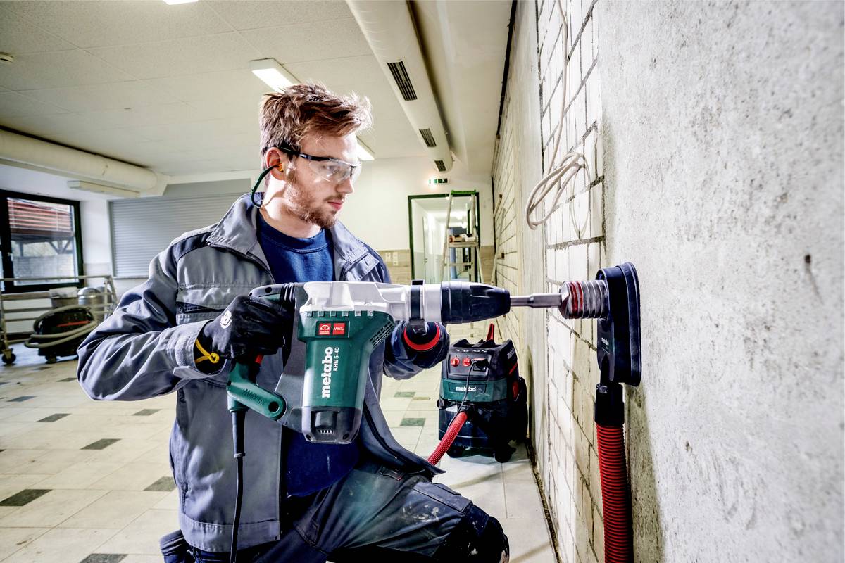 Ein Mann bearbeitet mit einem Bohrhammer eine Betonwand. Er trägt Schutzbrille und Gehörschutz, in einer Werkstattumgebung.
