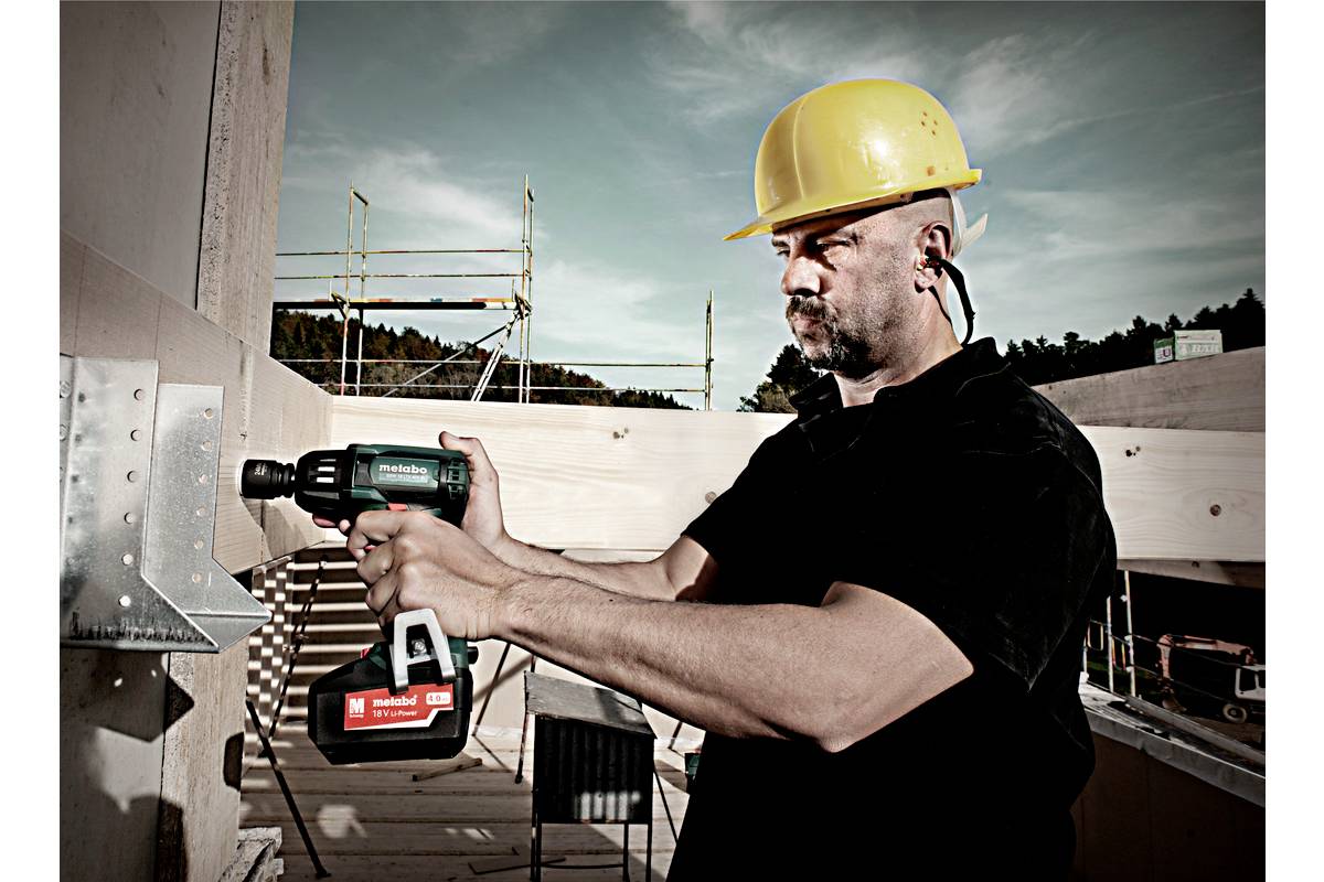 Ein Bauarbeiter mit gelbem Schutzhelm schraubt mit einem Akkuschrauber an einer Holzstruktur auf einer Baustelle im Freien.