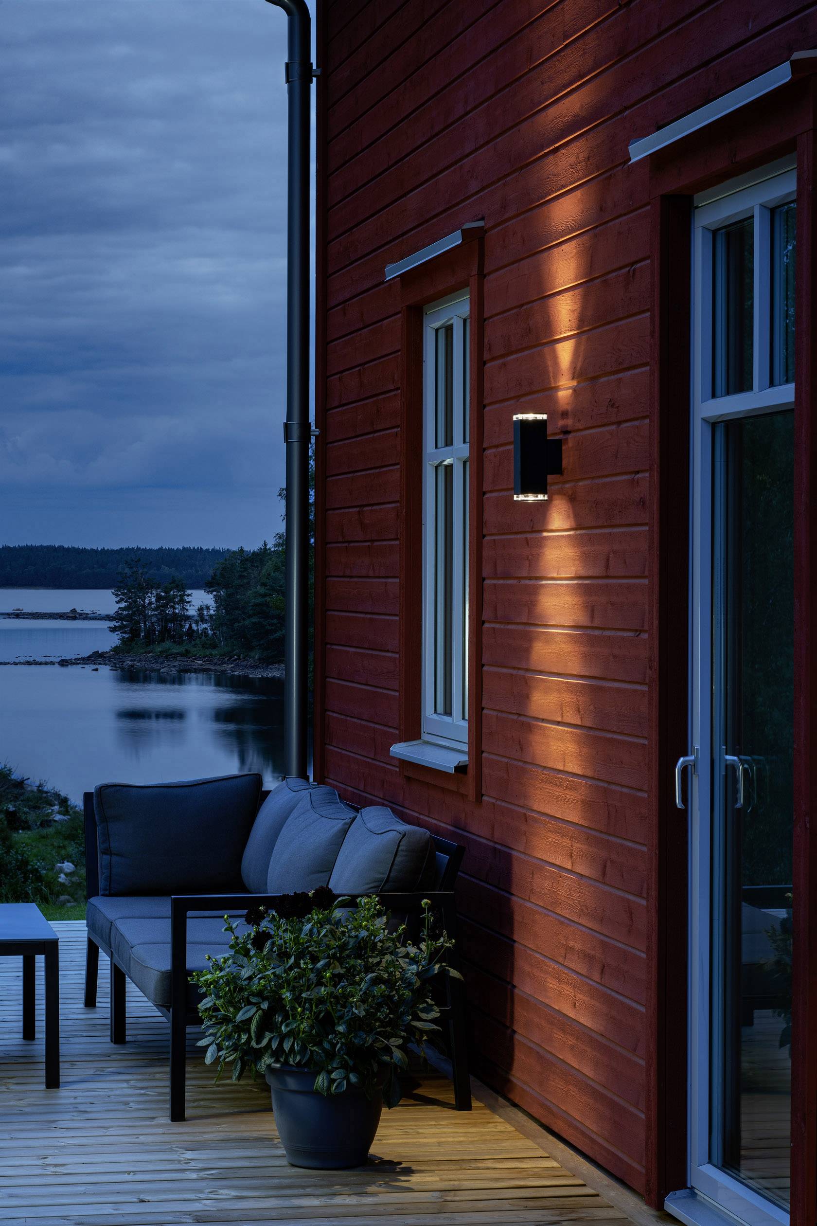 Rotes Haus am See bei Dämmerung, Terrasse mit Sofa und Pflanze, ruhige Atmosphäre, Wasser und bewaldete Küste im Hintergrund.