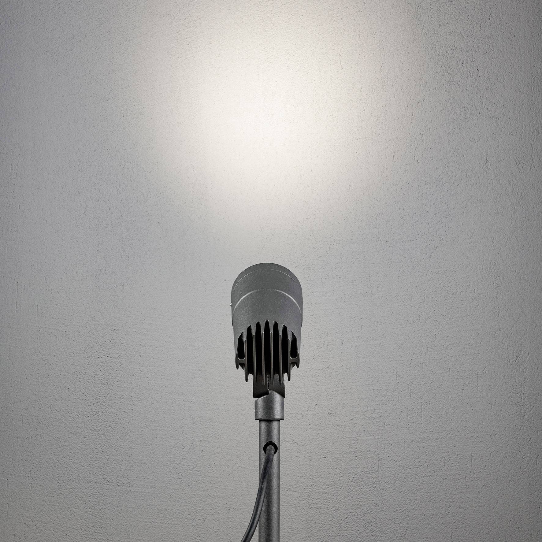 Ein Mikrofon steht im Vordergrund vor einer leeren Wand, von oben beleuchtet, symbolisiert bereit für eine Ansprache oder Präsentation.