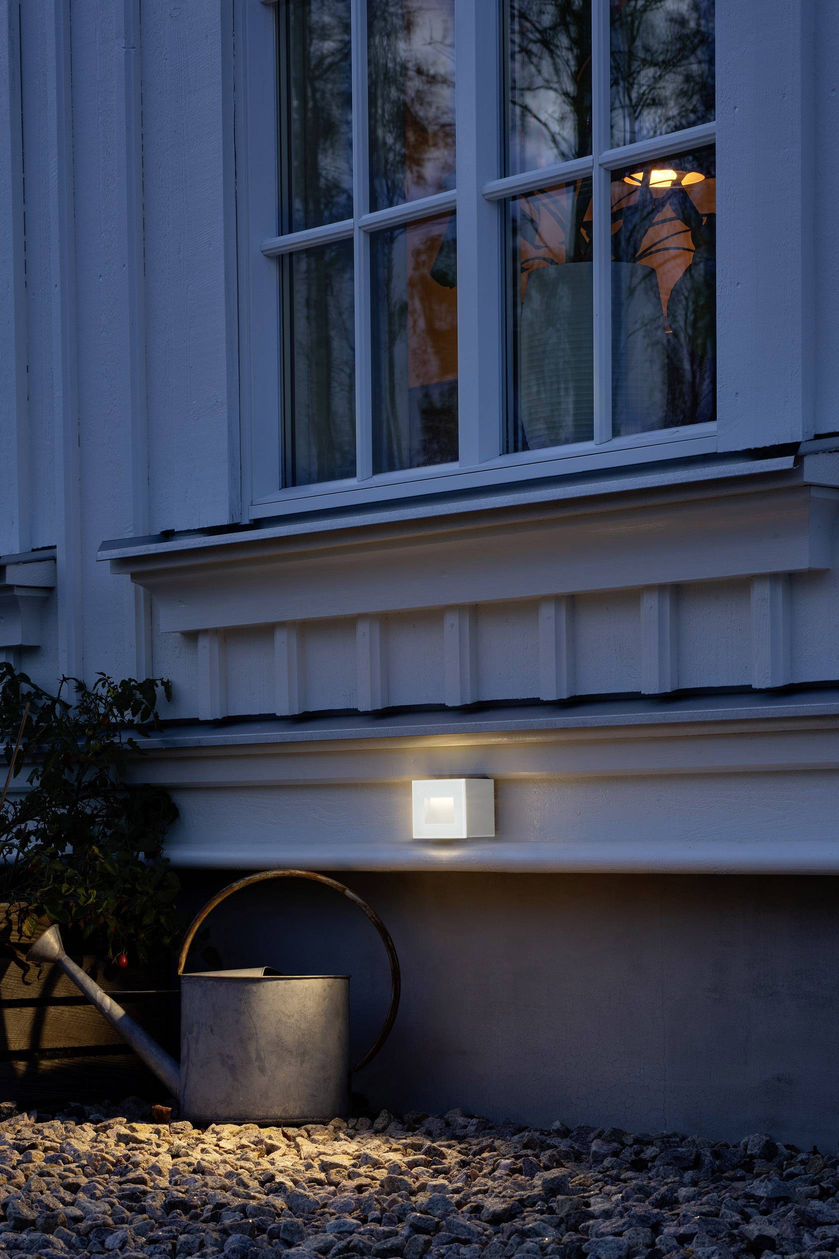 Wandleuchte leuchtet an einer Hausfassade neben einem Fenster. Darunter Kiesboden mit Gießkanne. Dämmerung verstärkt die Beleuchtungseffekte.