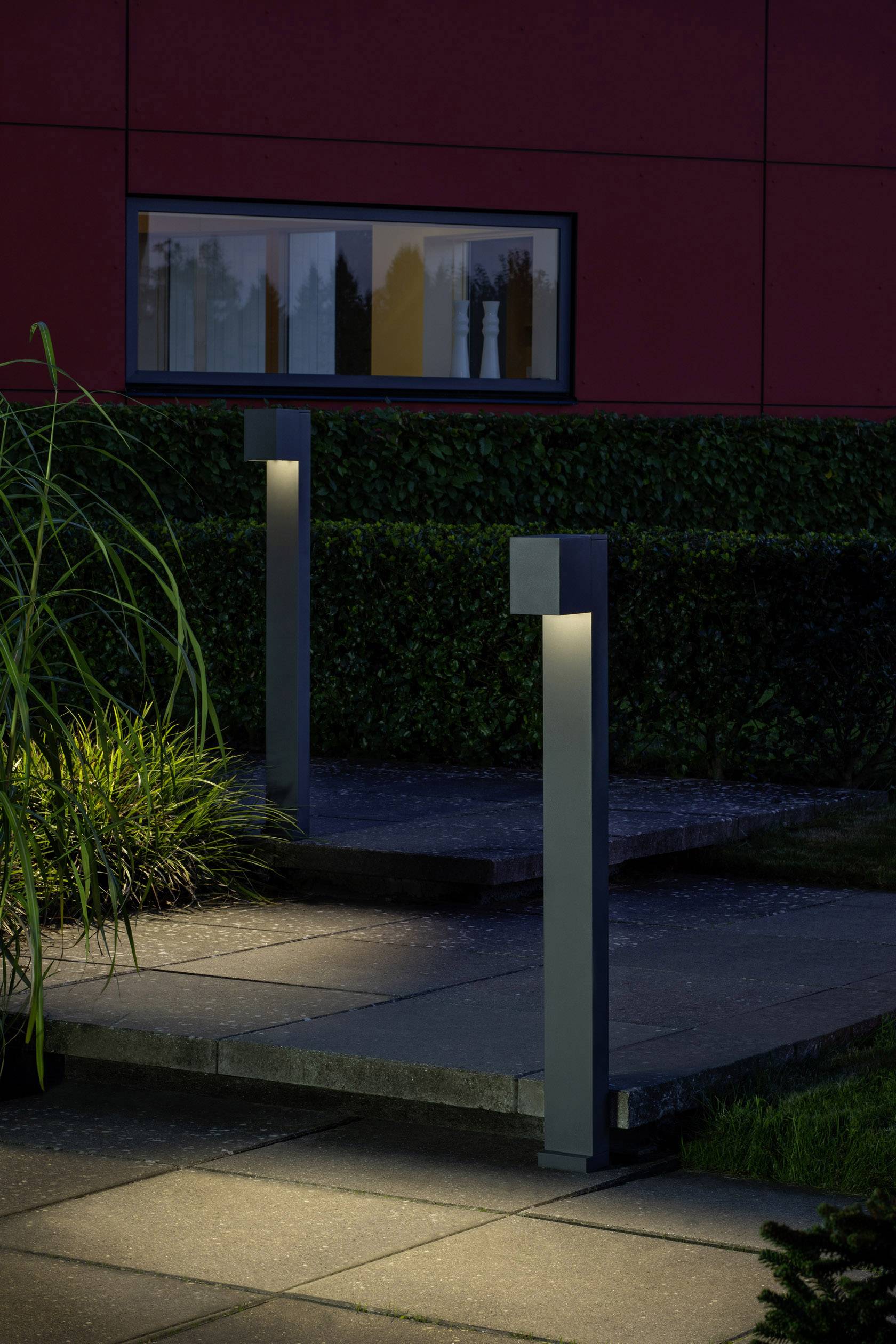 Gartenweg mit modernen, leuchtenden Stehlampen neben einem roten Gebäude und grünem Laubwerk.