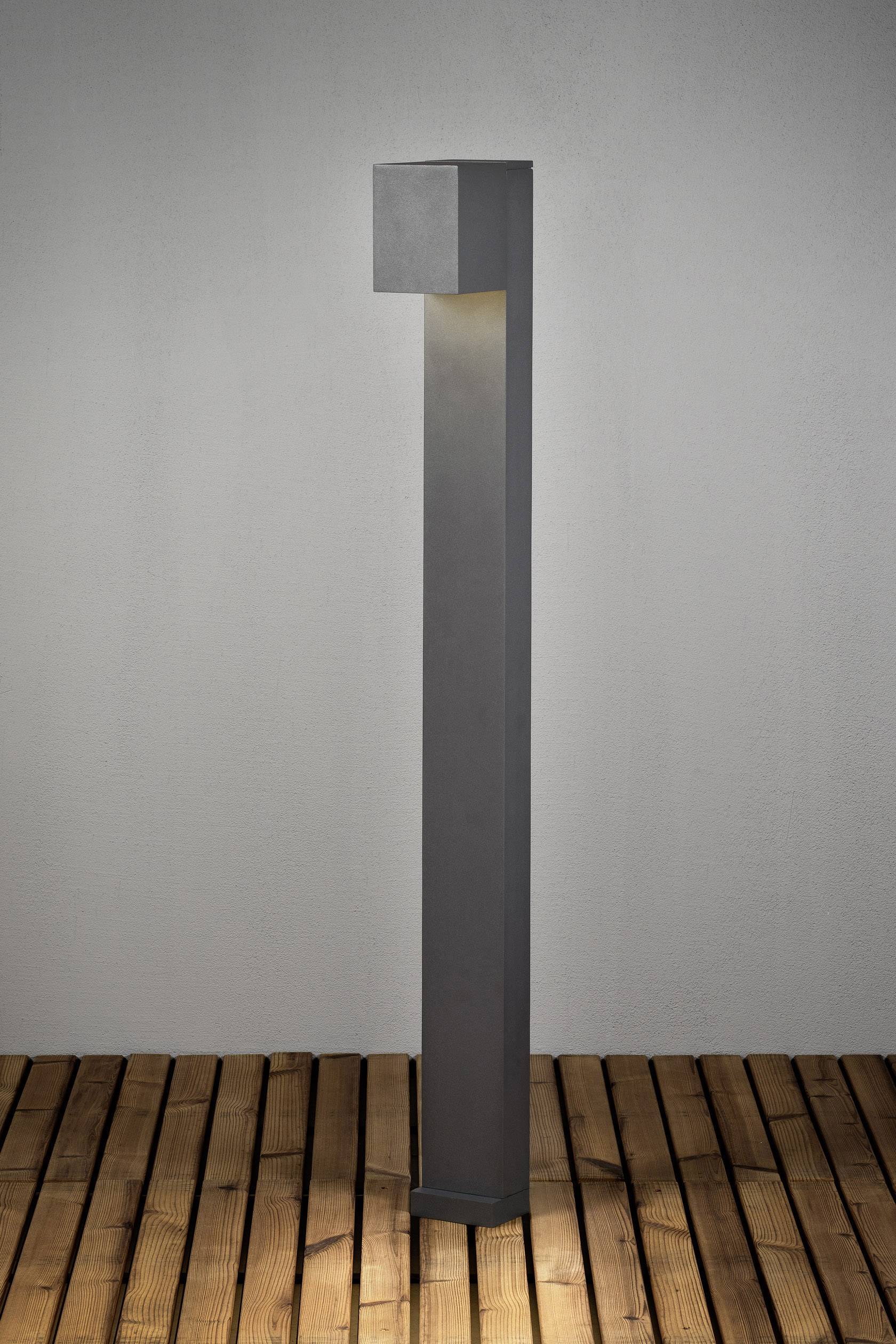 Eine moderne, rechteckige Außenleuchte aus Metall steht auf einem Holzdeck. Sie leuchtet nach unten und erzeugt ein warmes Licht.