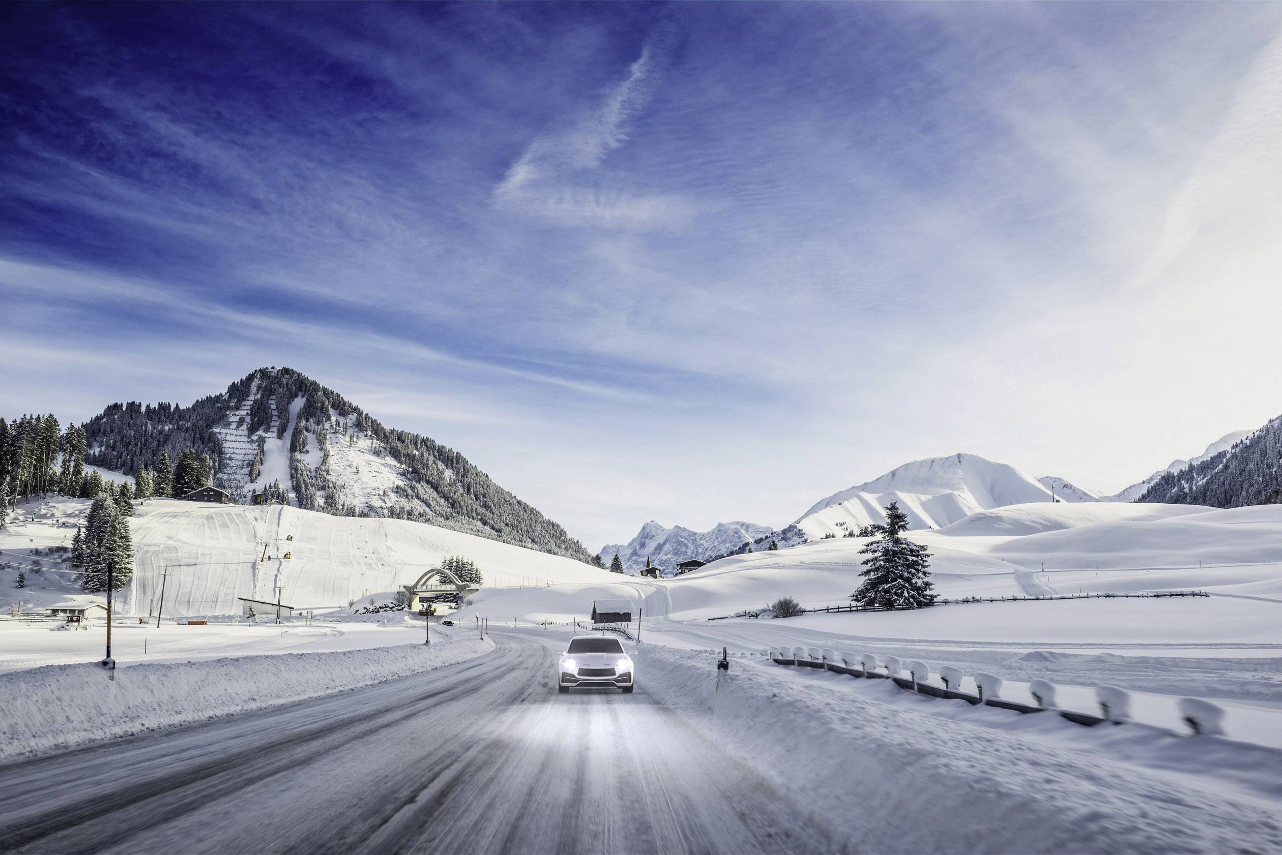 Ein Auto fährt auf einer verschneiten Straße durch eine winterliche Berglandschaft mit klarem Himmel und schneebedeckten Bergen im Hintergrund.