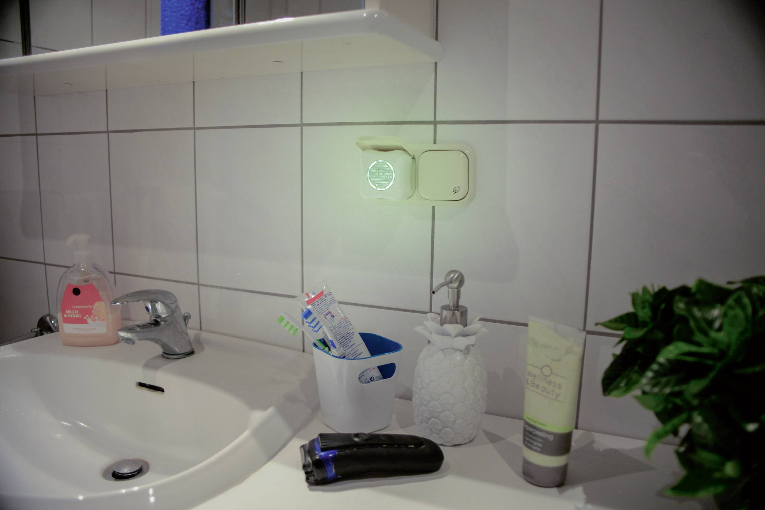 Ein Badezimmer mit einem Waschbecken, Zahnpasta, Seifenspender und Rasierer. An der Wand ist ein beleuchteter Schalter angebracht.