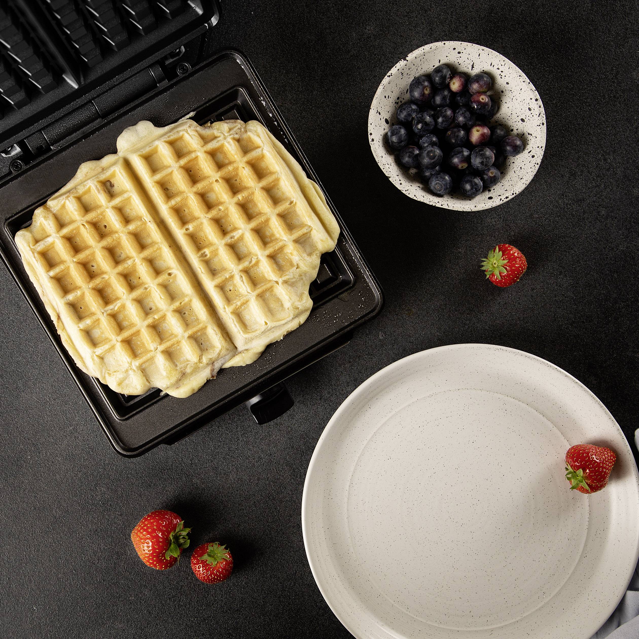 Belgischer Waffeleisen mit frischen Waffeln, daneben Schüssel mit Blaubeeren und Erdbeeren auf schwarzem Hintergrund.