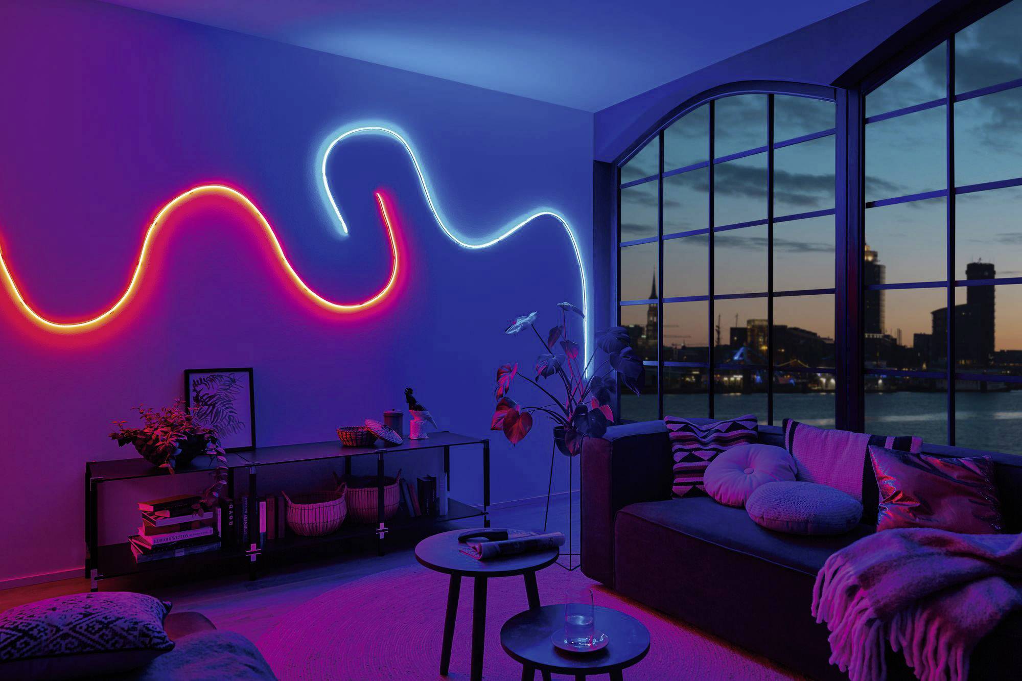 Wohnzimmer mit moderner Beleuchtung: An der Wand leuchtet ein geschwungener LED-Streifen in Rot und Blau. Große Fenster zeigen eine nächtliche Stadtansicht.
