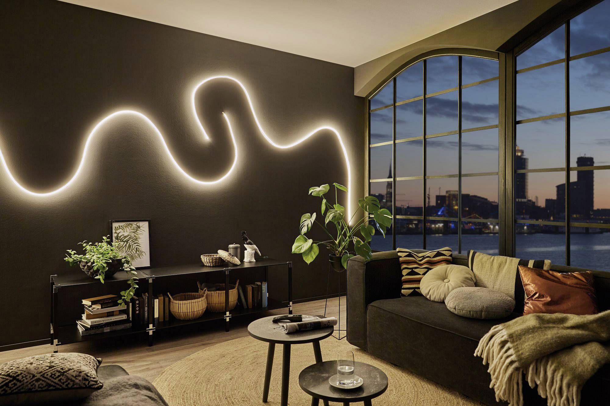 Wohnzimmer mit modernem Design: Dunkle Wand mit leuchtendem LED-Streifen, große Fenster mit Blick auf Stadtlandschaft bei Sonnenuntergang, elegante schwarze Möbel, gemütliches Sofa.