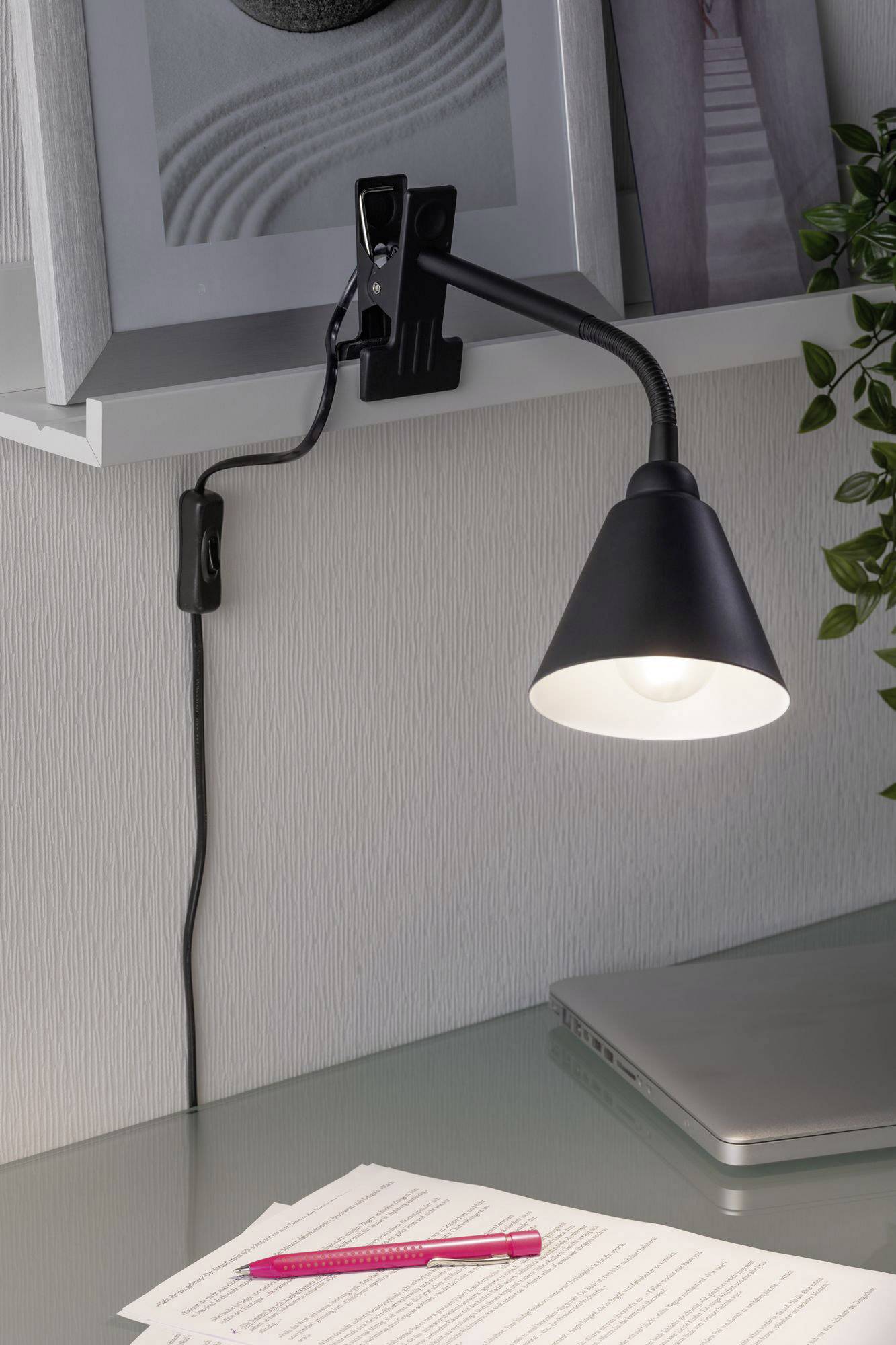 Eine schwarze Klemmleuchte leuchtet auf einen Schreibtisch, darauf liegen ein aufgeschlagenes Notizbuch, ein roter Stift und ein geschlossener Laptop.