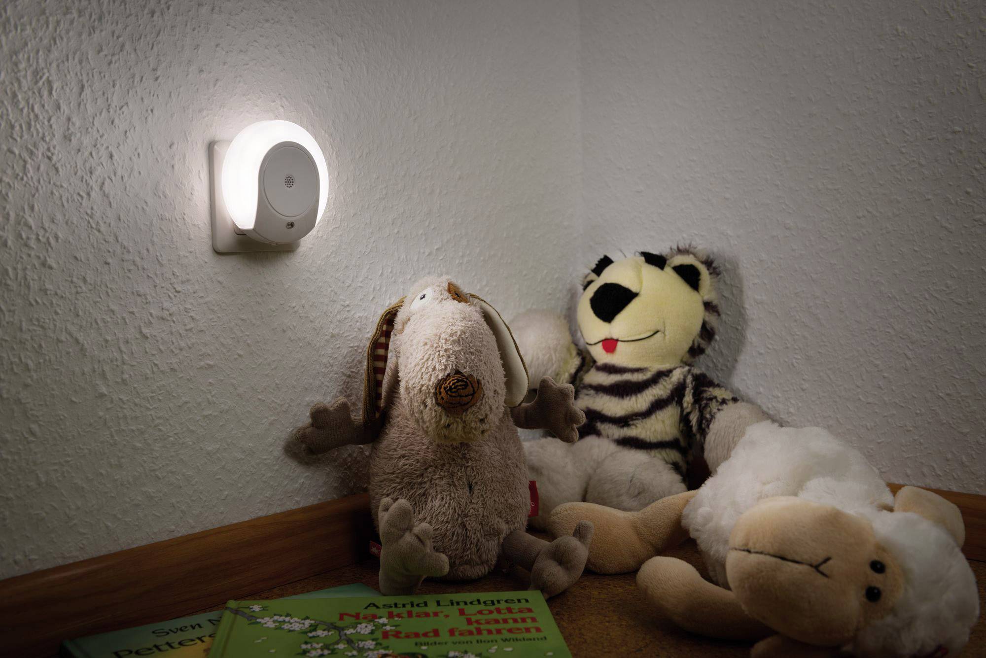 Eine Ecke eines Kinderzimmers mit einem Nachtlicht an der Wand. Darunter sitzen Stofftiere und Kinderbücher.