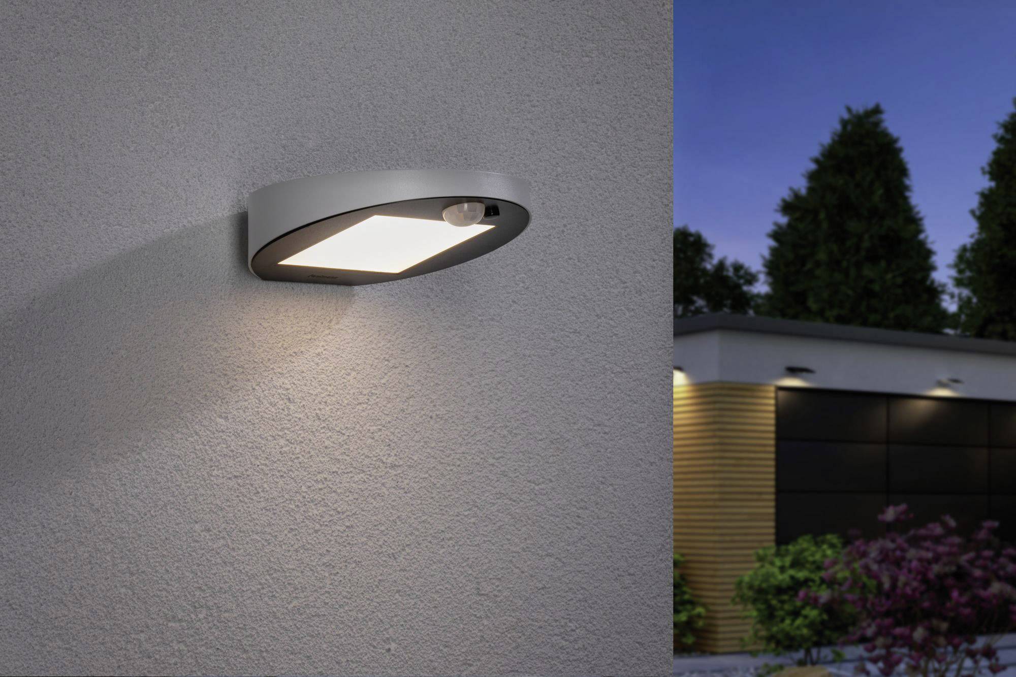 Außenlichtquelle mit rechteckiger Form an einer Wand, strahlt warmes Licht aus. Im Hintergrund ein modernes Gebäude in der Dämmerung.